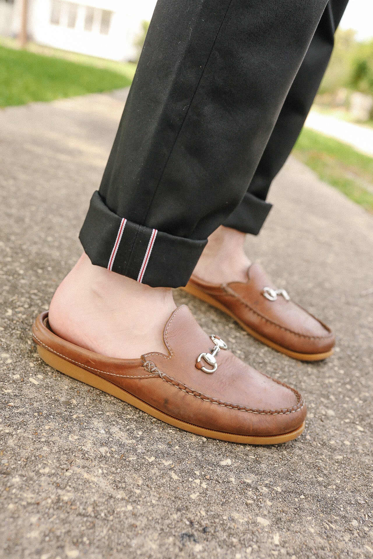 Close-up of feet wearing brown leather slip-on loafers with metal bit detail and black cuffed pants