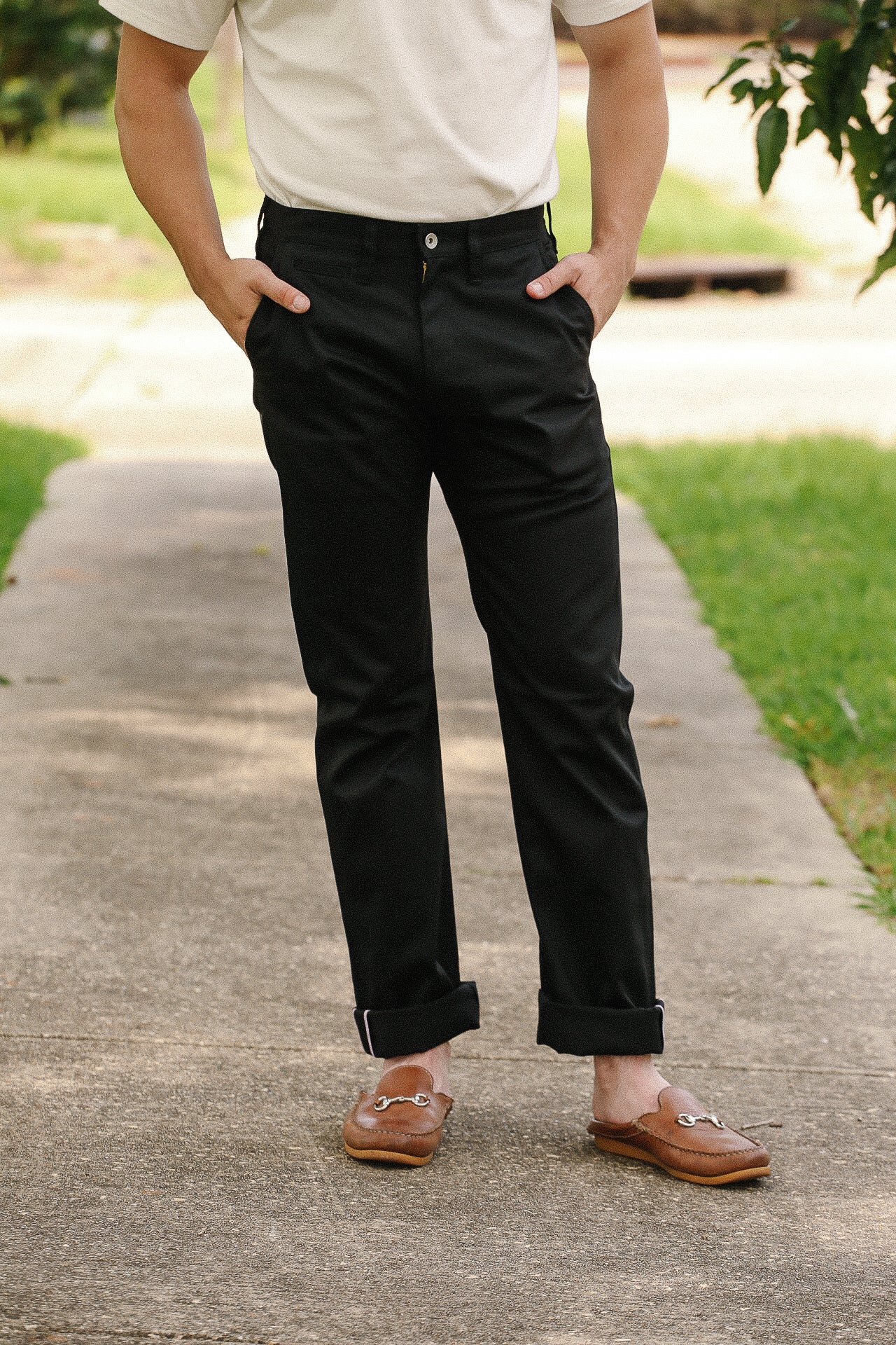 Man wearing black pants and brown slip-on leather loafers standing on a sidewalk with hands in pockets