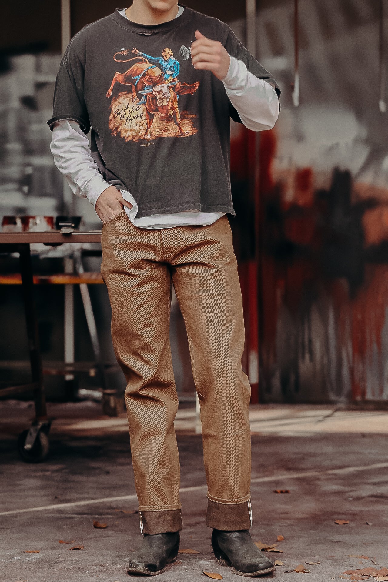Person wearing layered long-sleeve shirt with bull rider graphic and beige work pants with black boots in workshop