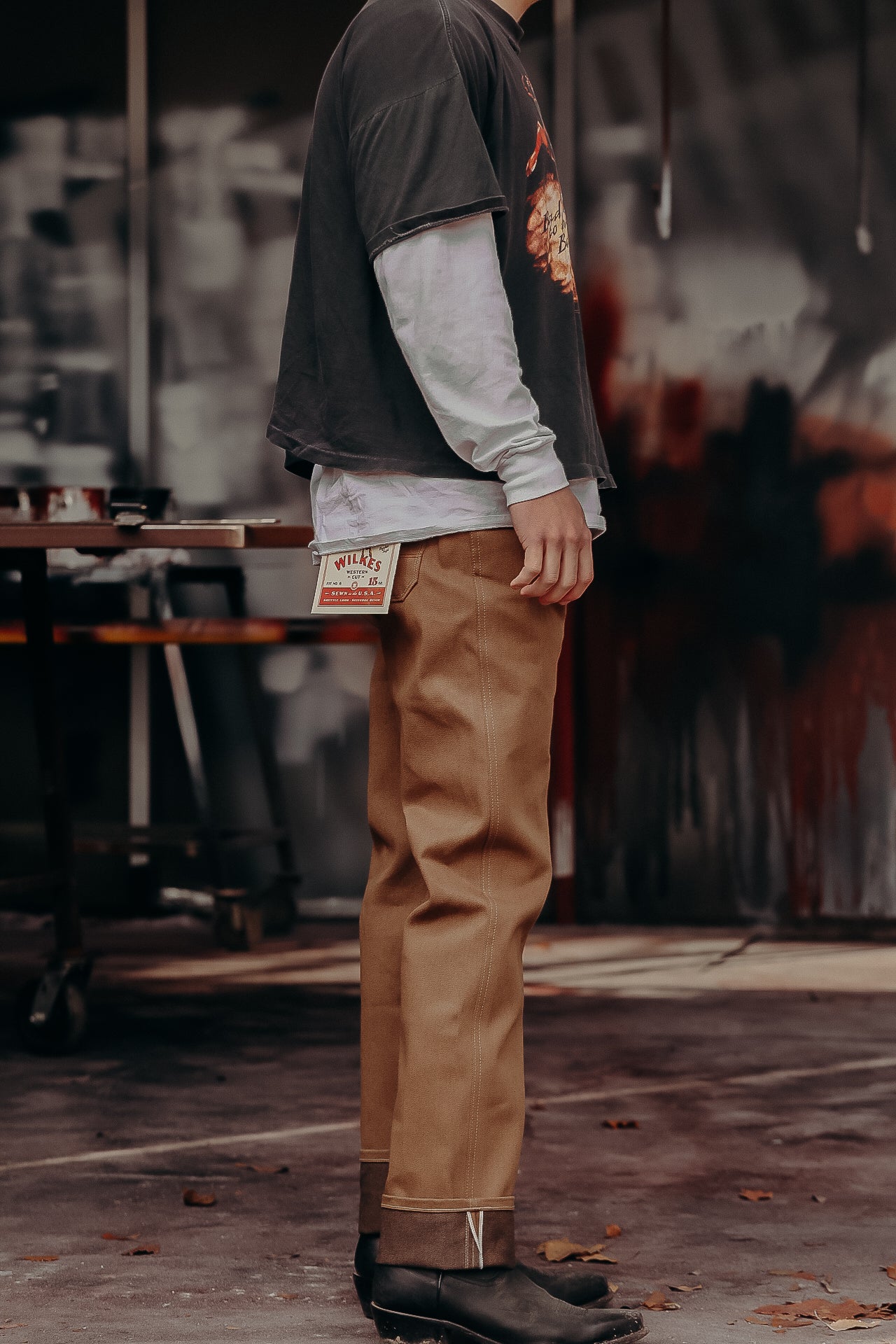 Person wearing Wilkes Western Cut tan work pants with a visible label, black boots, and layered long sleeve shirts in rustic shop setting