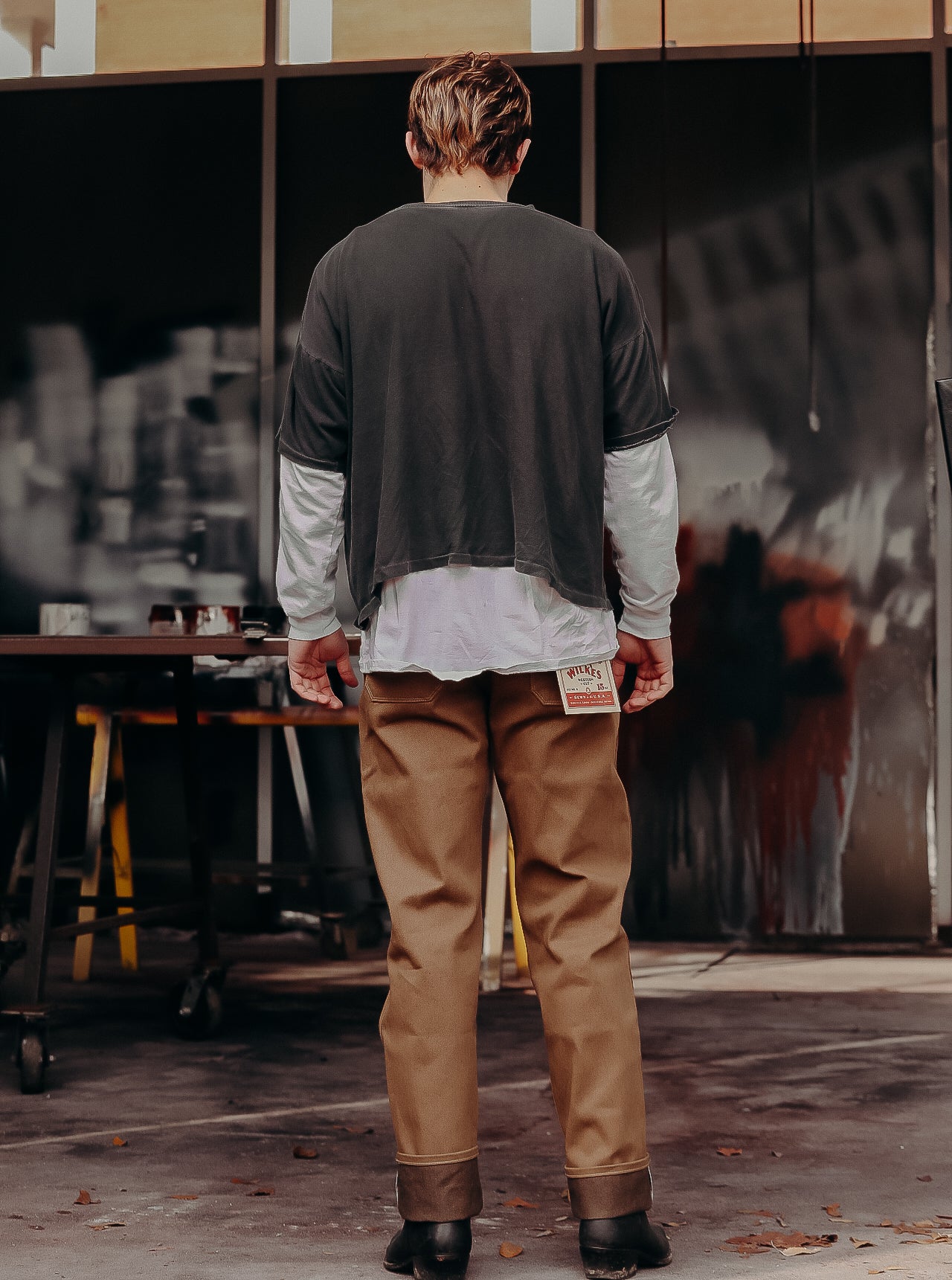 Man wearing brown work pants with label, layered shirts, and black boots in workshop setting at Iron Shop Provisions
