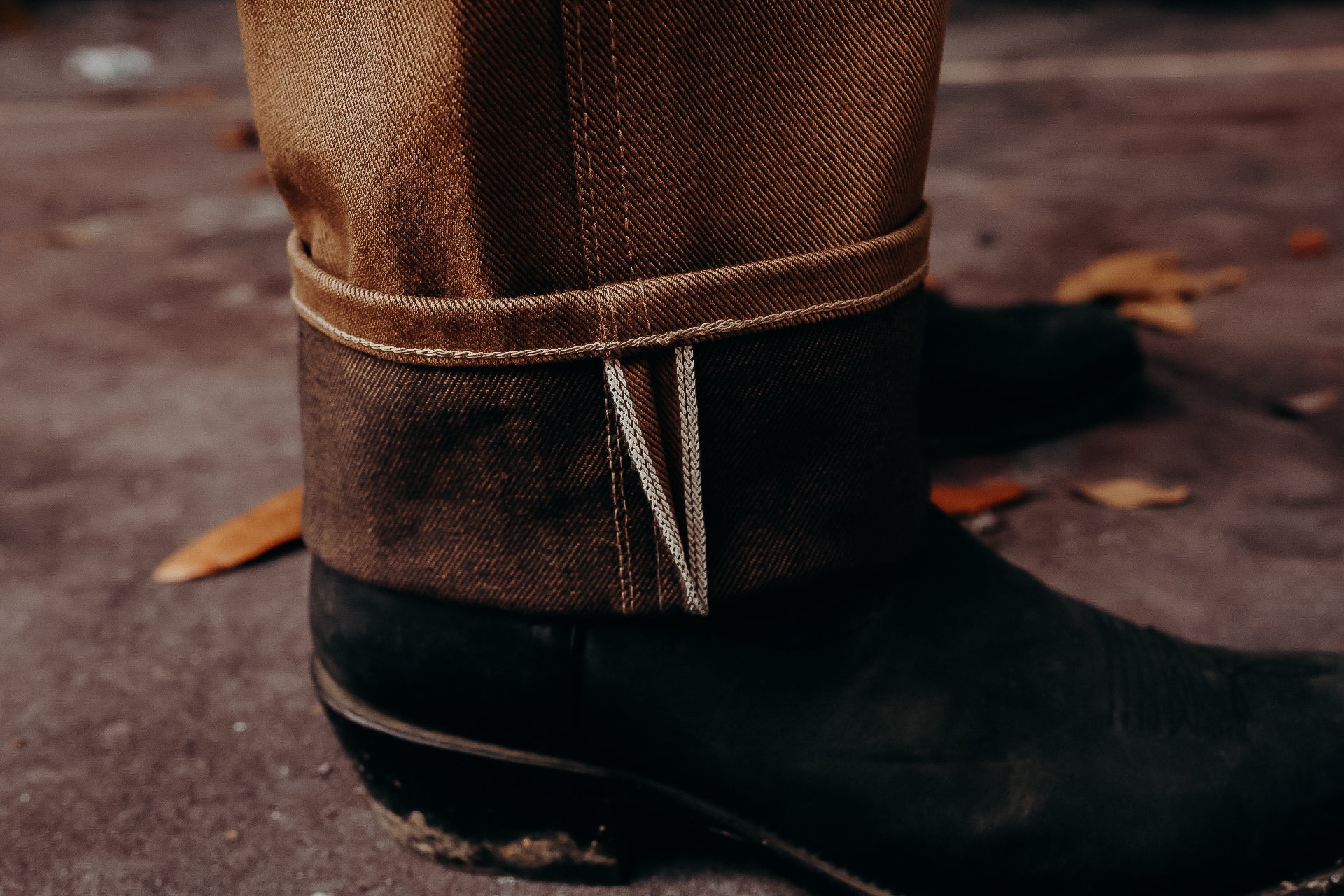Close-up of brown selvedge denim jeans cuffed above black leather boots on concrete floor with autumn leaves