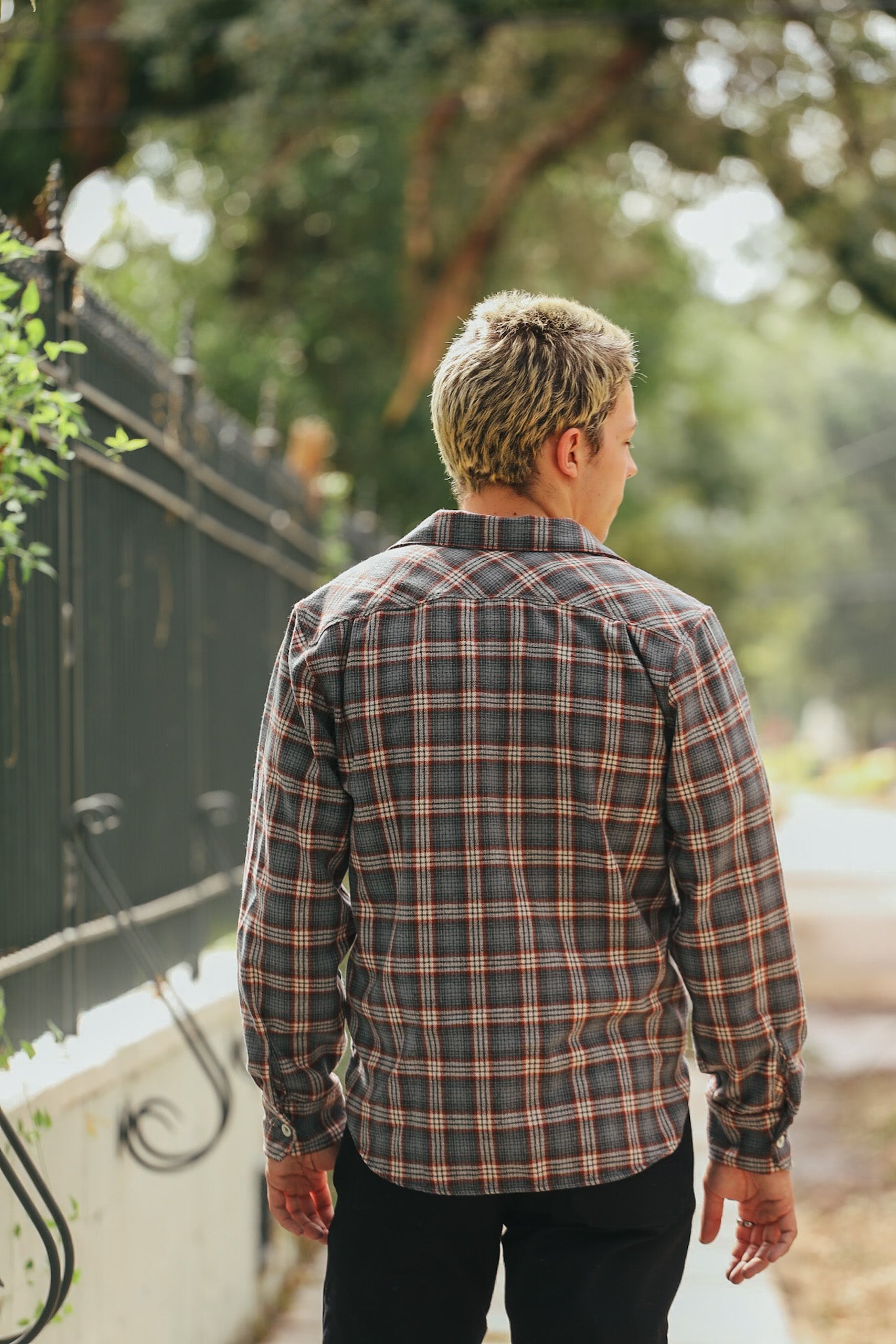 Man wearing a gray and red plaid flannel shirt standing outdoors by a black iron fence