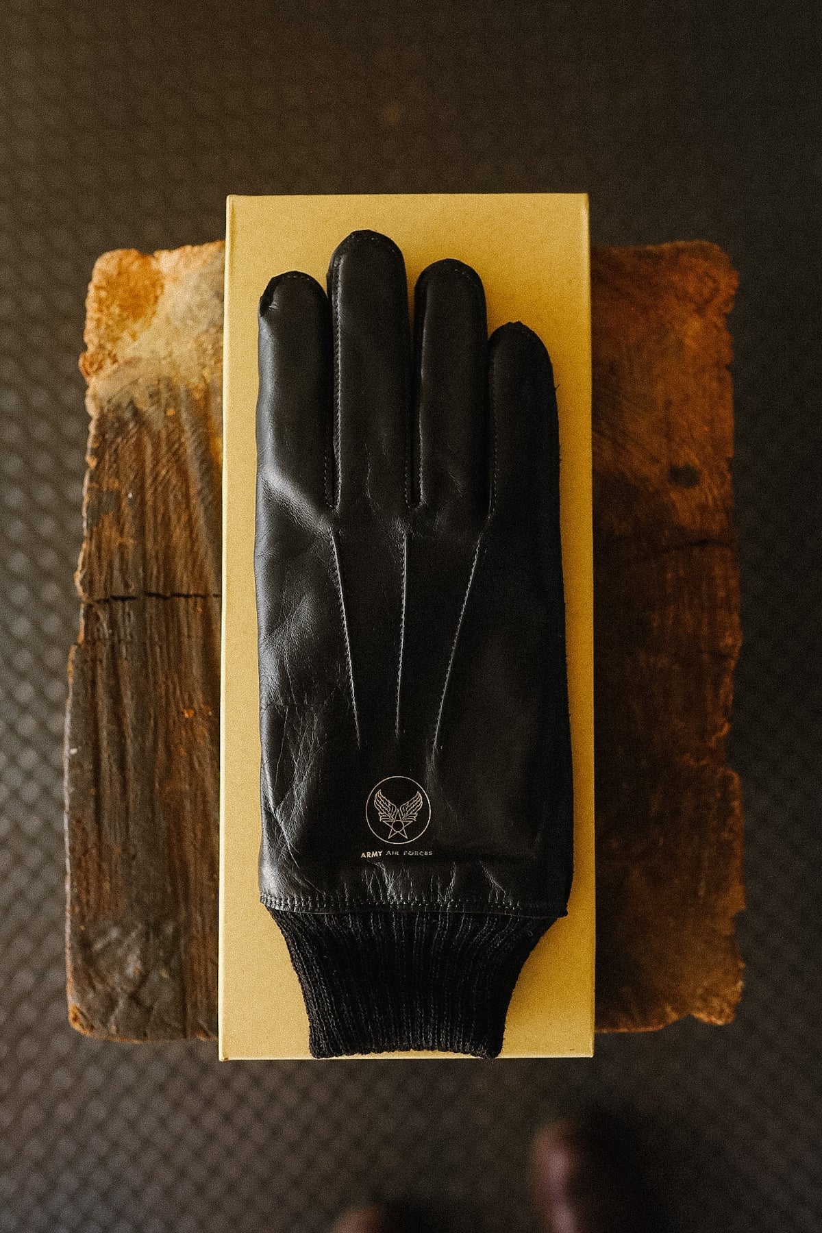A black leather glove with a ribbed knit cuff, displayed on top of a gold-colored rectangular box. The box is placed on two rustic wooden blocks over a dark textured floor.