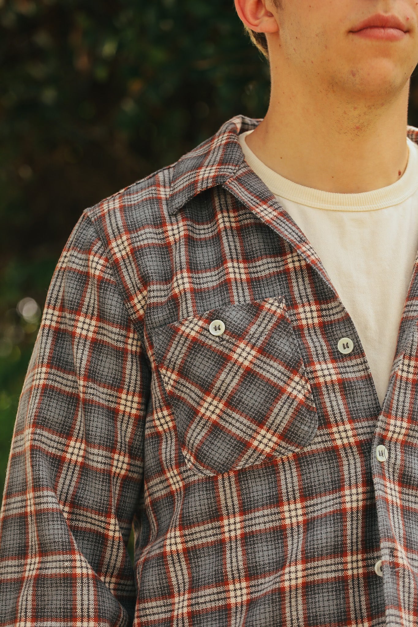 Close-up of a man wearing a gray and red plaid shirt over a cream-colored T-shirt, outdoor background