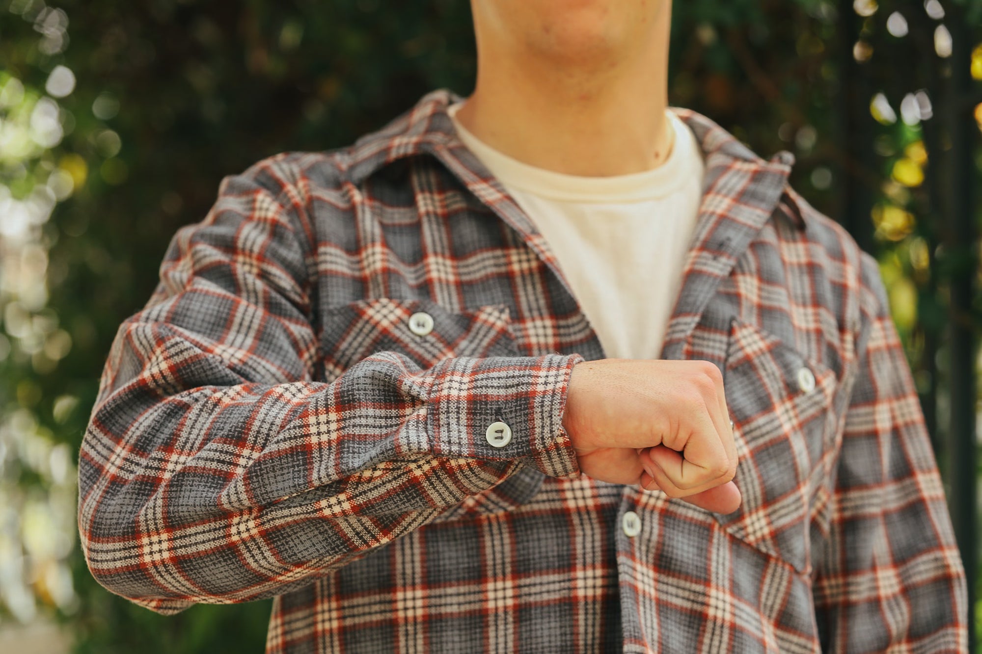 Man wearing a gray and orange plaid shirt over white tee outdoors with trees in background
