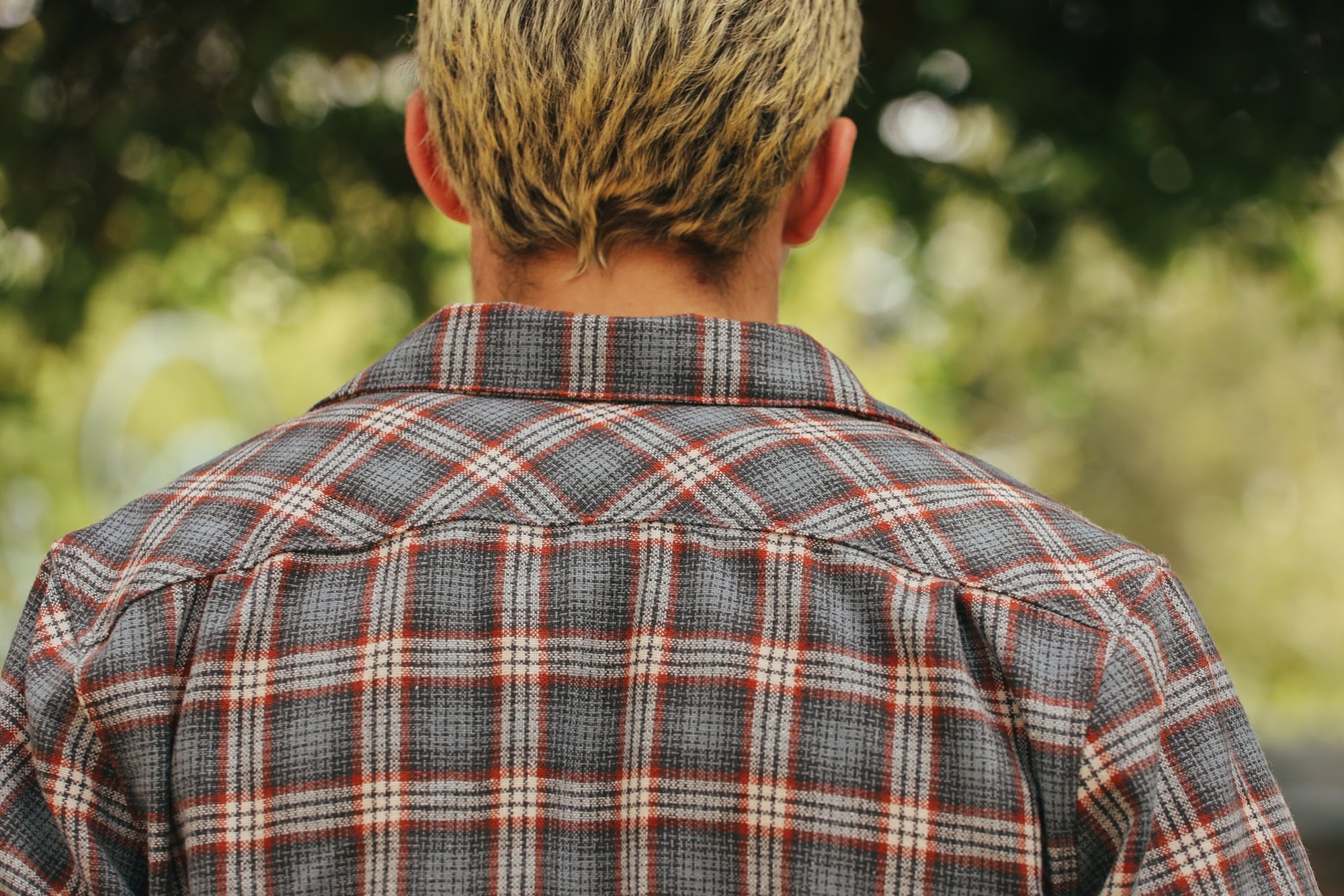Back view of person with blonde hair wearing a gray and orange plaid shirt outdoors with green leafy background