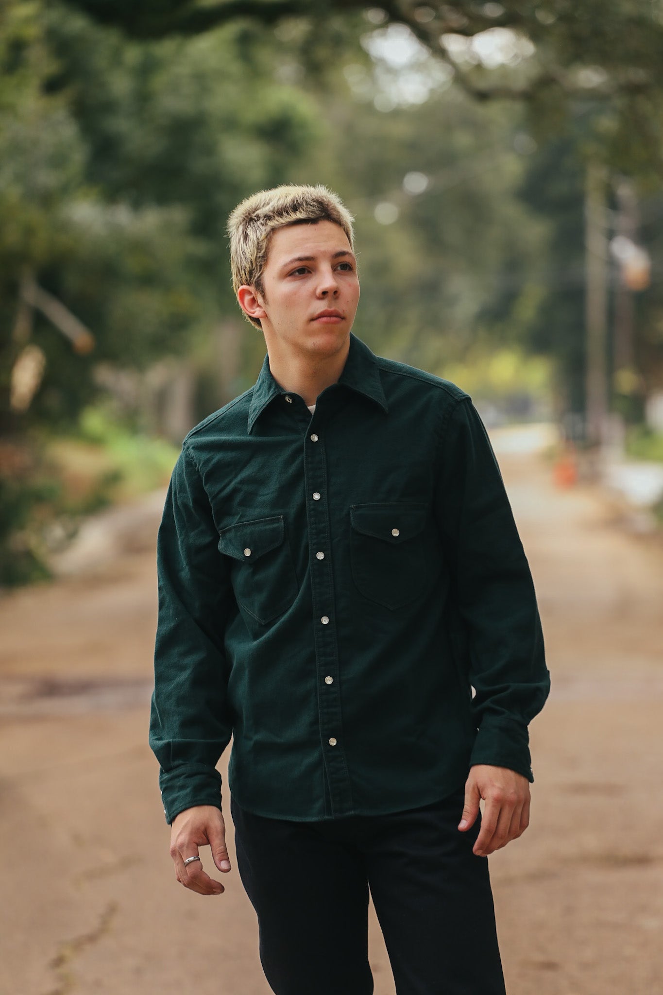 Young man wearing dark green button-up shirt standing outdoors on a rustic path with blurred trees