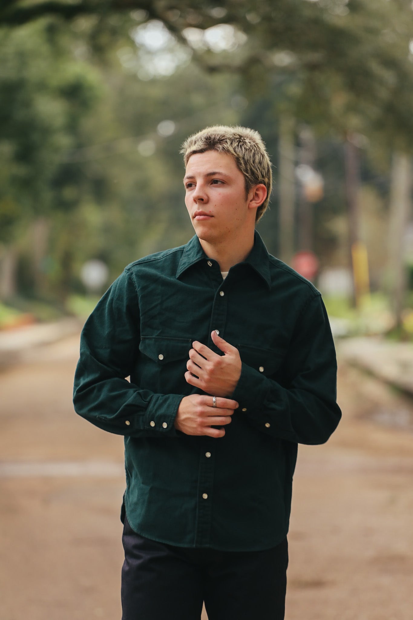 Young man wearing dark green button-up shirt standing outdoors on a tree-lined street