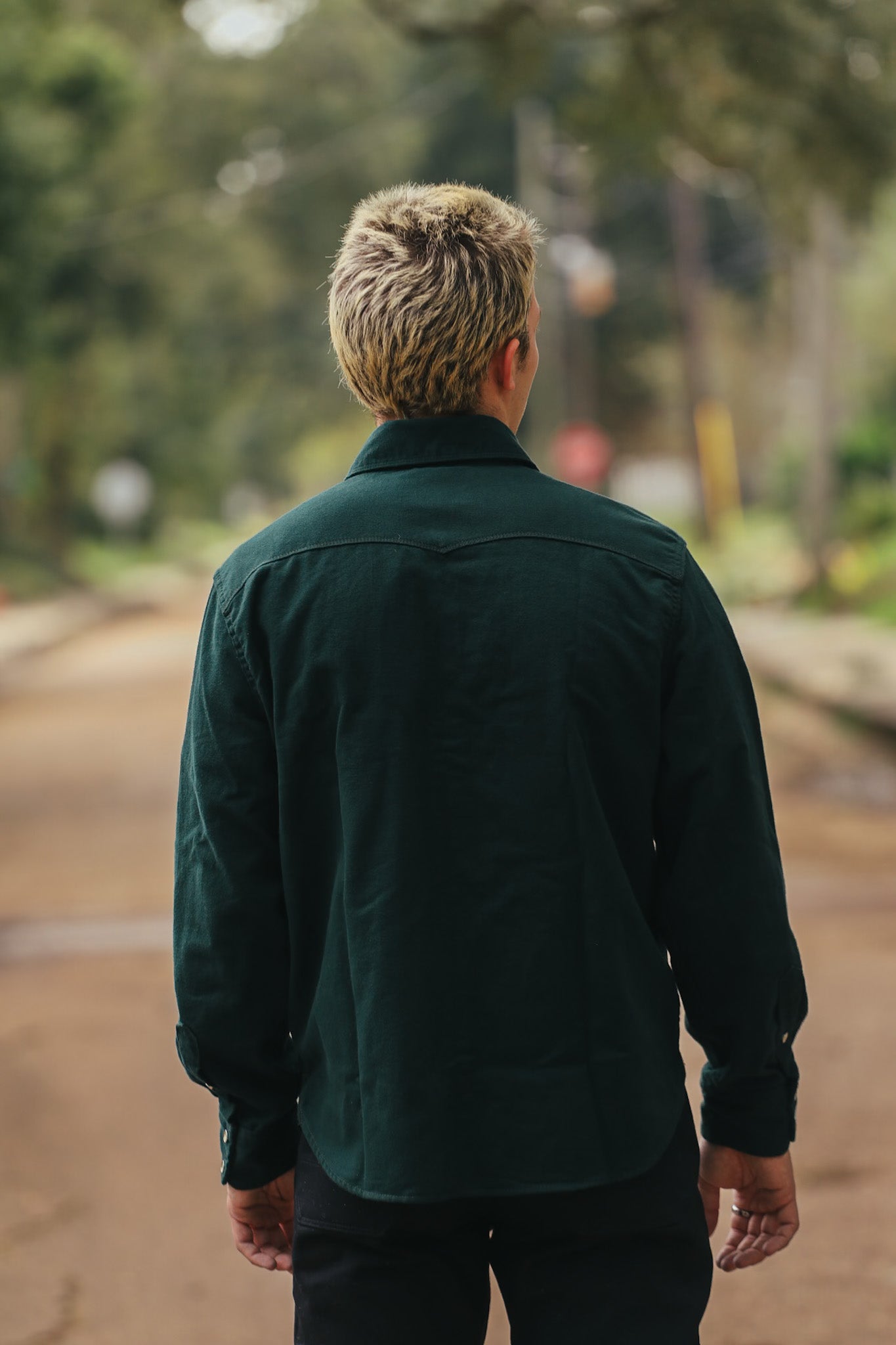 Man with blonde hair wearing dark green shirt standing on outdoor path with blurred nature background