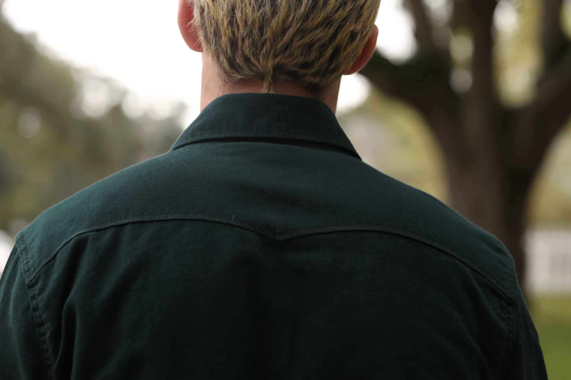 Man wearing dark green button-up shirt standing outdoors with blurred trees in background