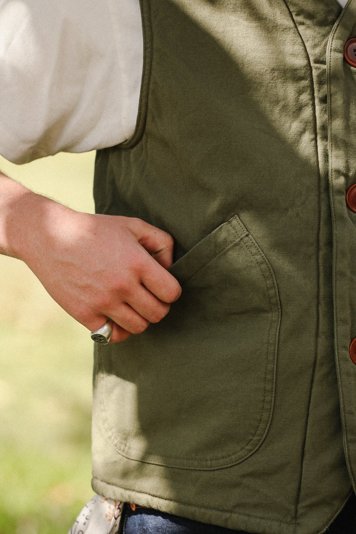 Close-up of a person wearing an olive green utility vest over a white shirt, with a hand reaching into a front pocket. The vest has brown buttons and is shown outdoors with a blurred green background.