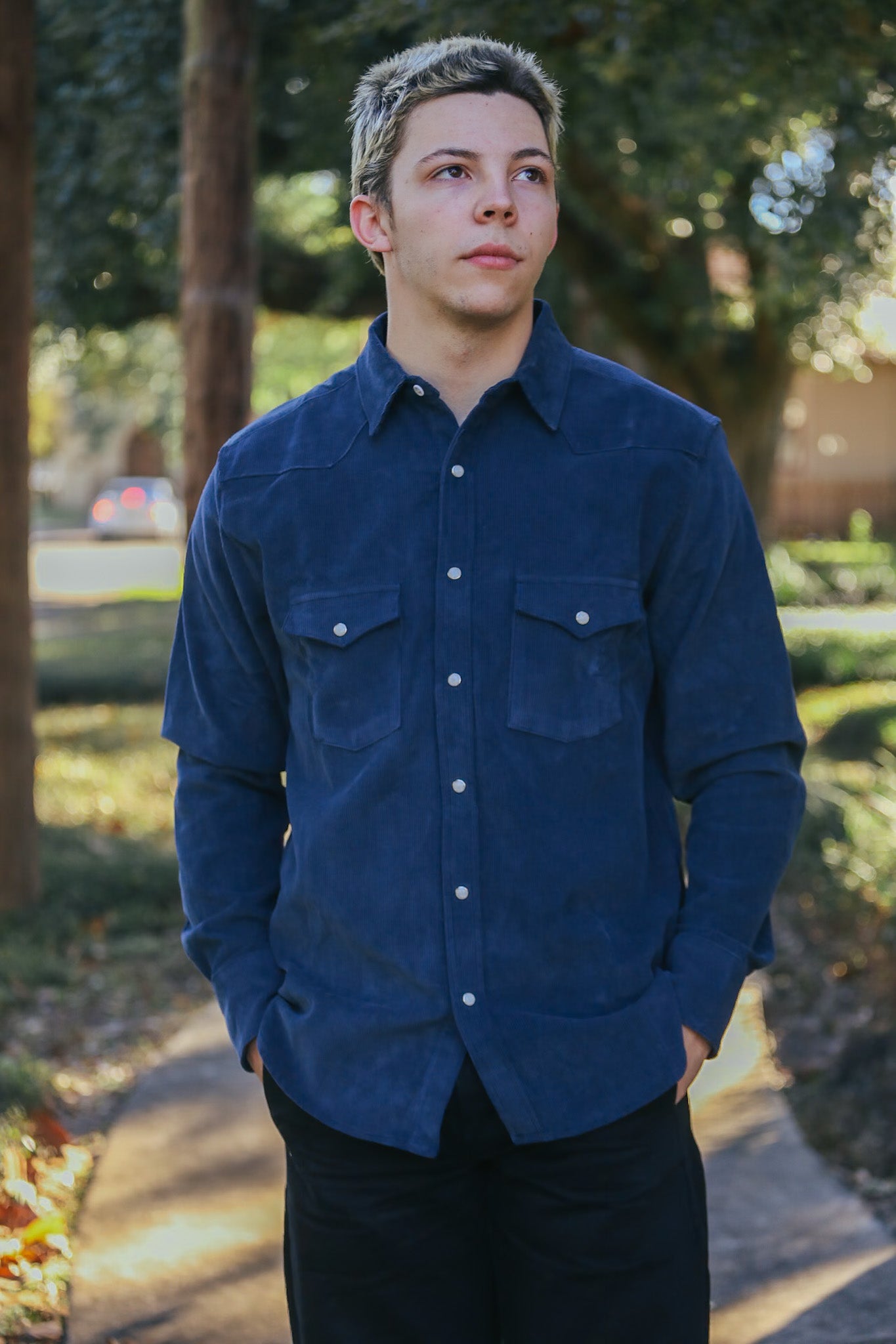 Young man wearing navy blue button-up shirt standing on outdoor path with trees in background