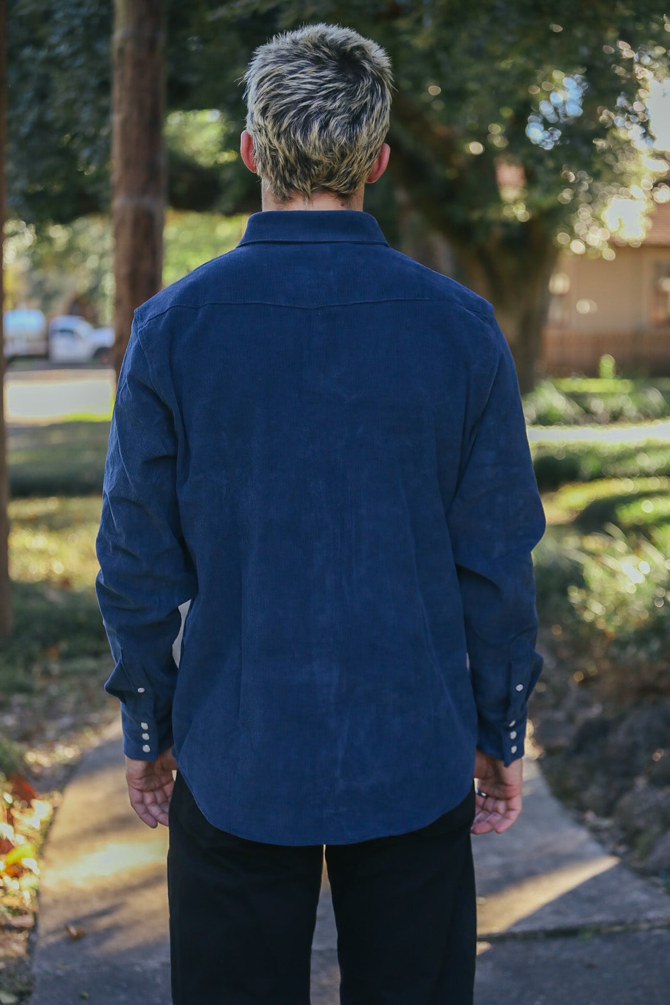 Man wearing navy blue corduroy long-sleeve shirt and black pants standing outdoors on a sunny path