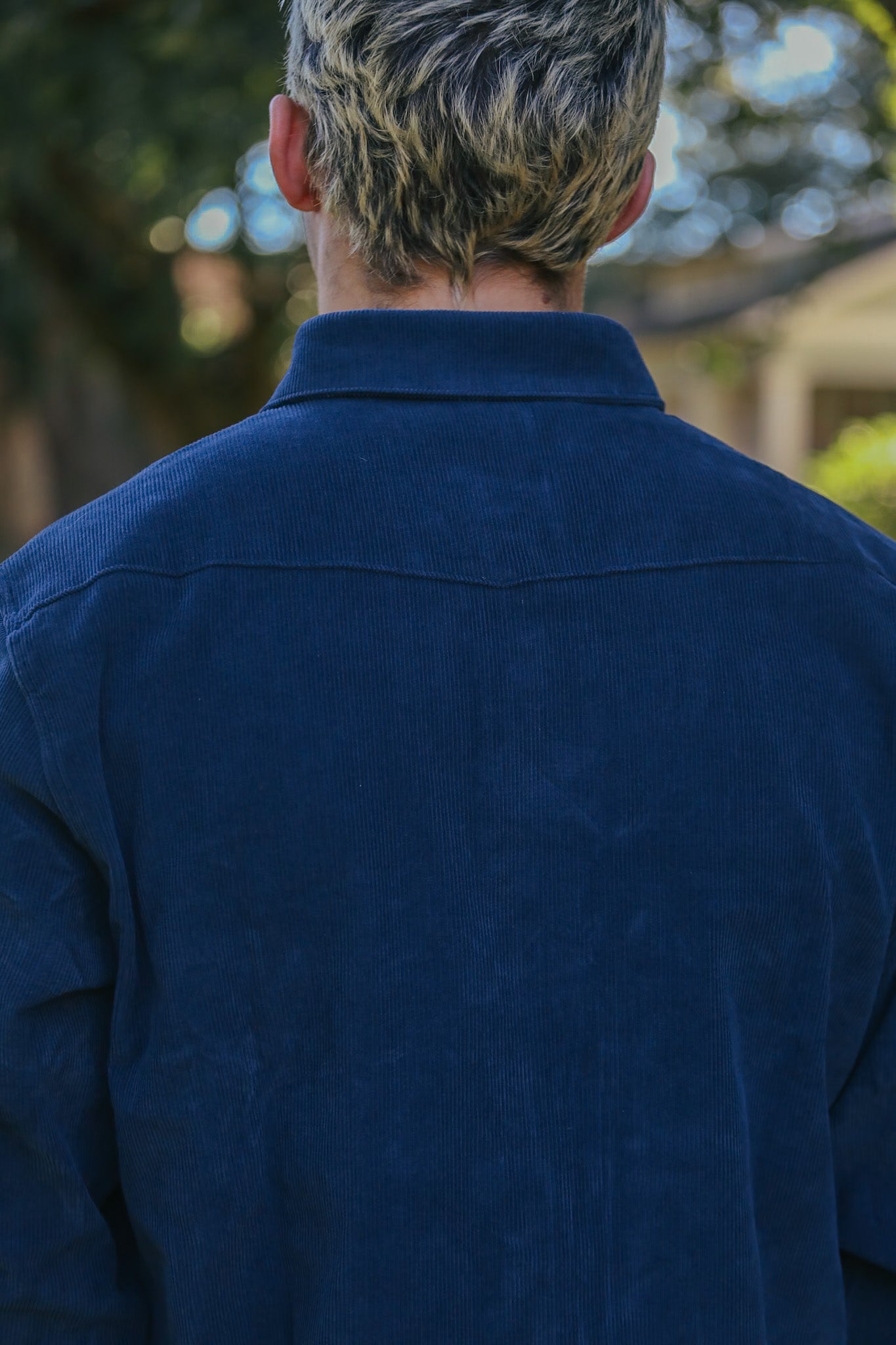 Back view of person wearing a dark blue corduroy shirt outdoors with blurred trees and house background