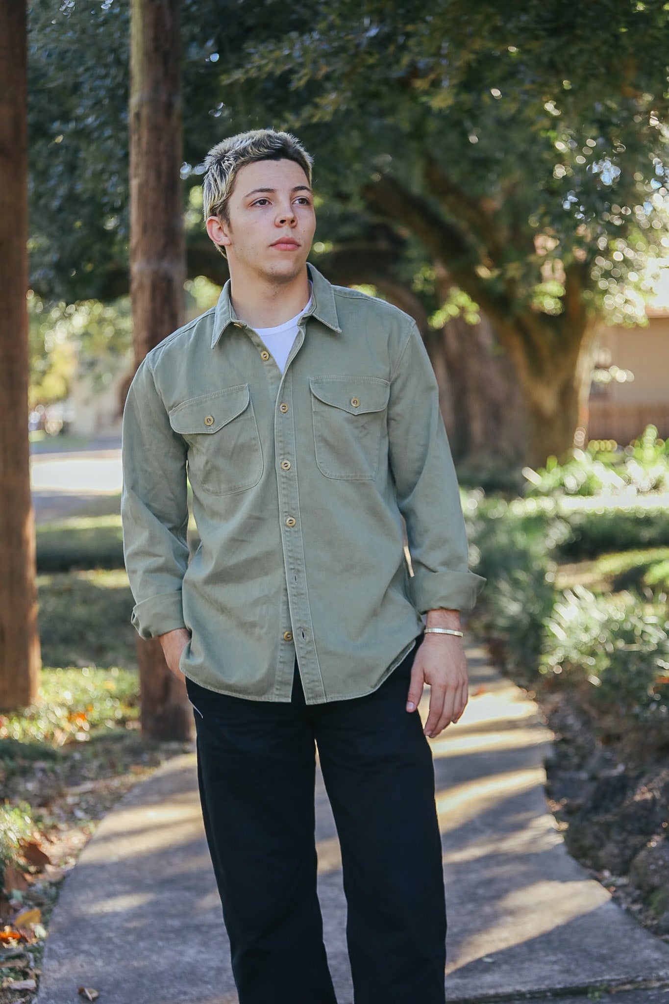Young man wearing olive green button-up work shirt over white tee and black pants walking on sunlit sidewalk