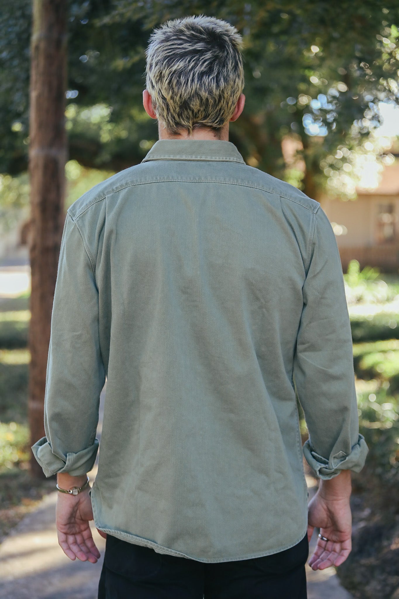 Person wearing olive green long-sleeve shirt with rolled cuffs standing outdoors with trees in background