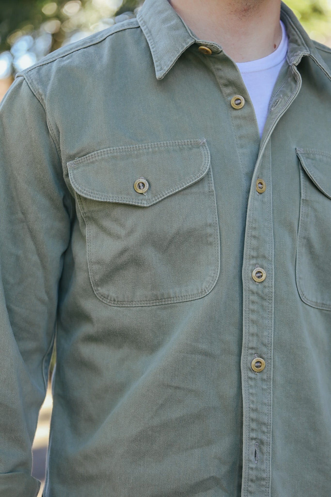 Close-up of a person wearing an olive green button-up shirt with flap chest pockets and a white undershirt outdoors