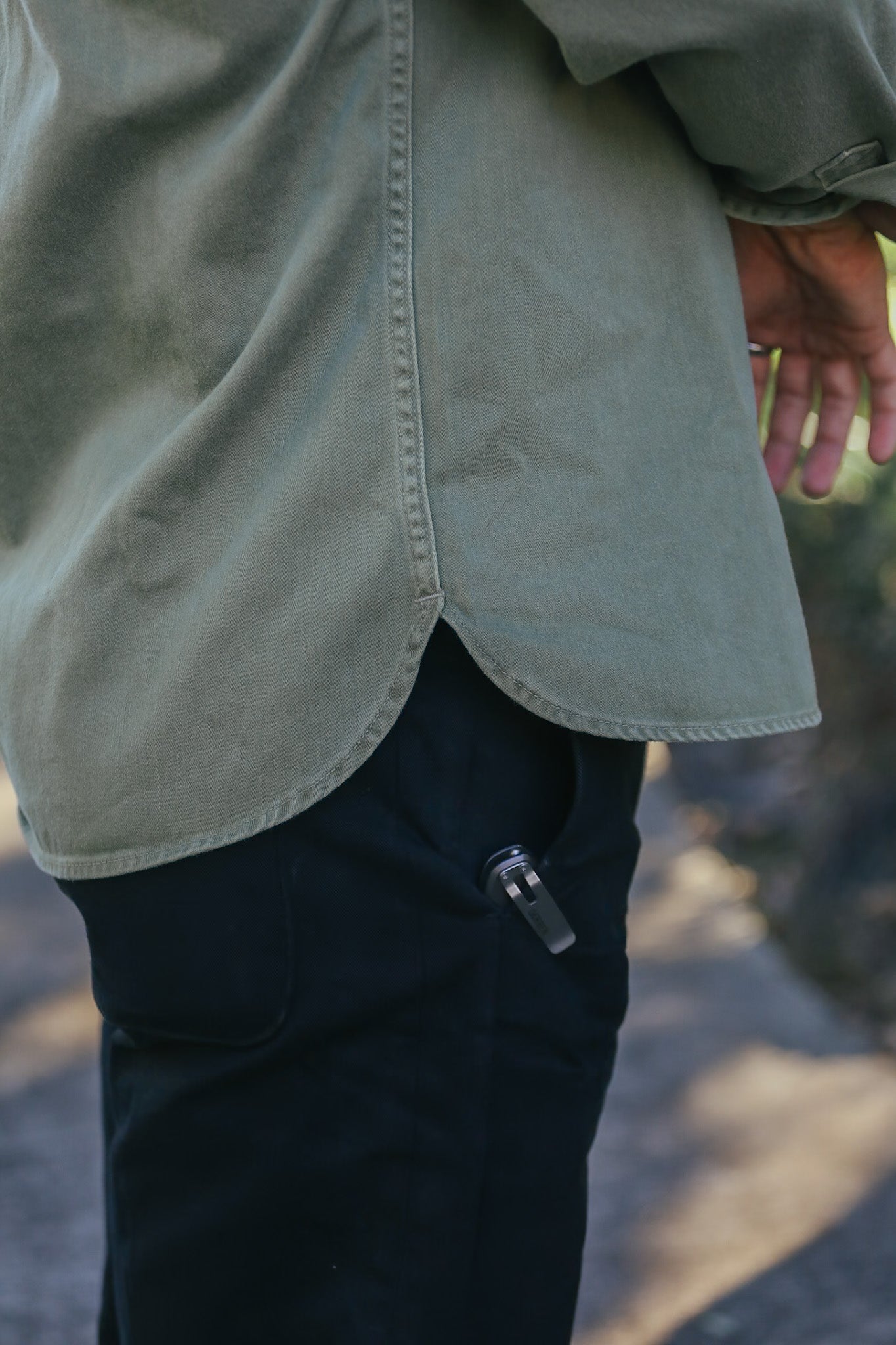 Person wearing olive green jacket and black pants with metal tool clipped to pocket outdoors