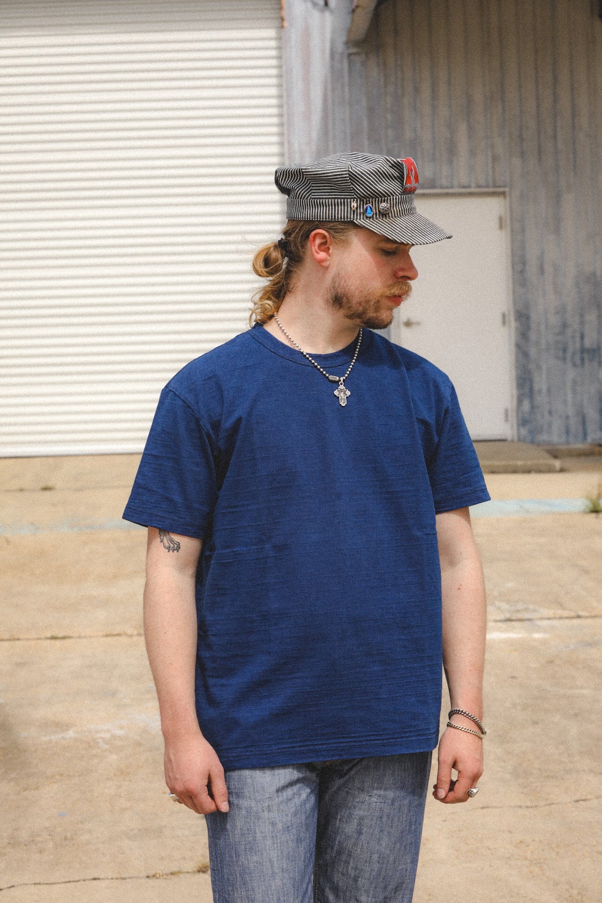 A man wearing a dark blue crewneck t-shirt, blue jeans, and a gray cap with a red logo, standing outdoors in front of a garage door and wooden building.