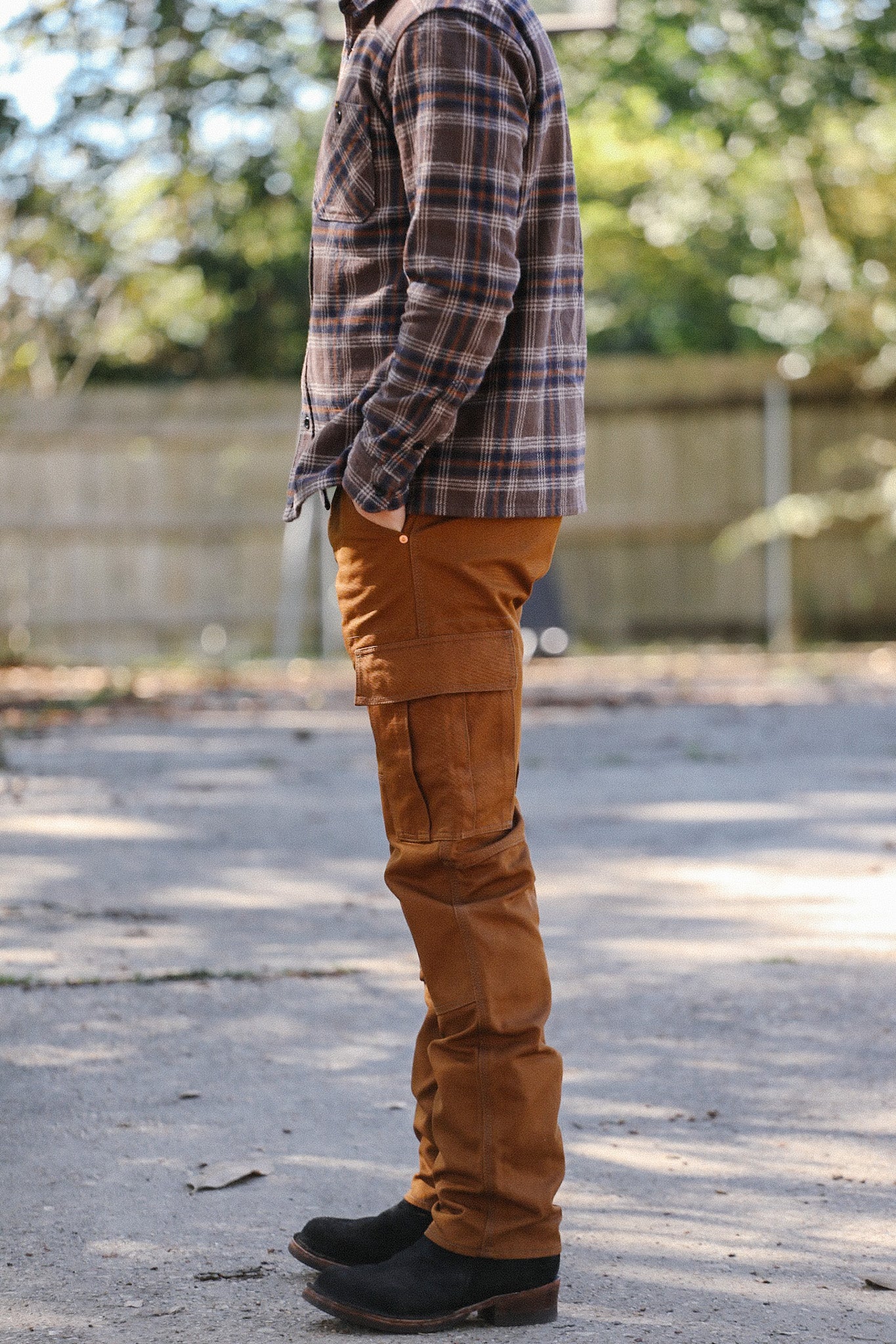 Side view of a man wearing a brown plaid shirt, copper work pants with reinforced knees, and black boots outdoors