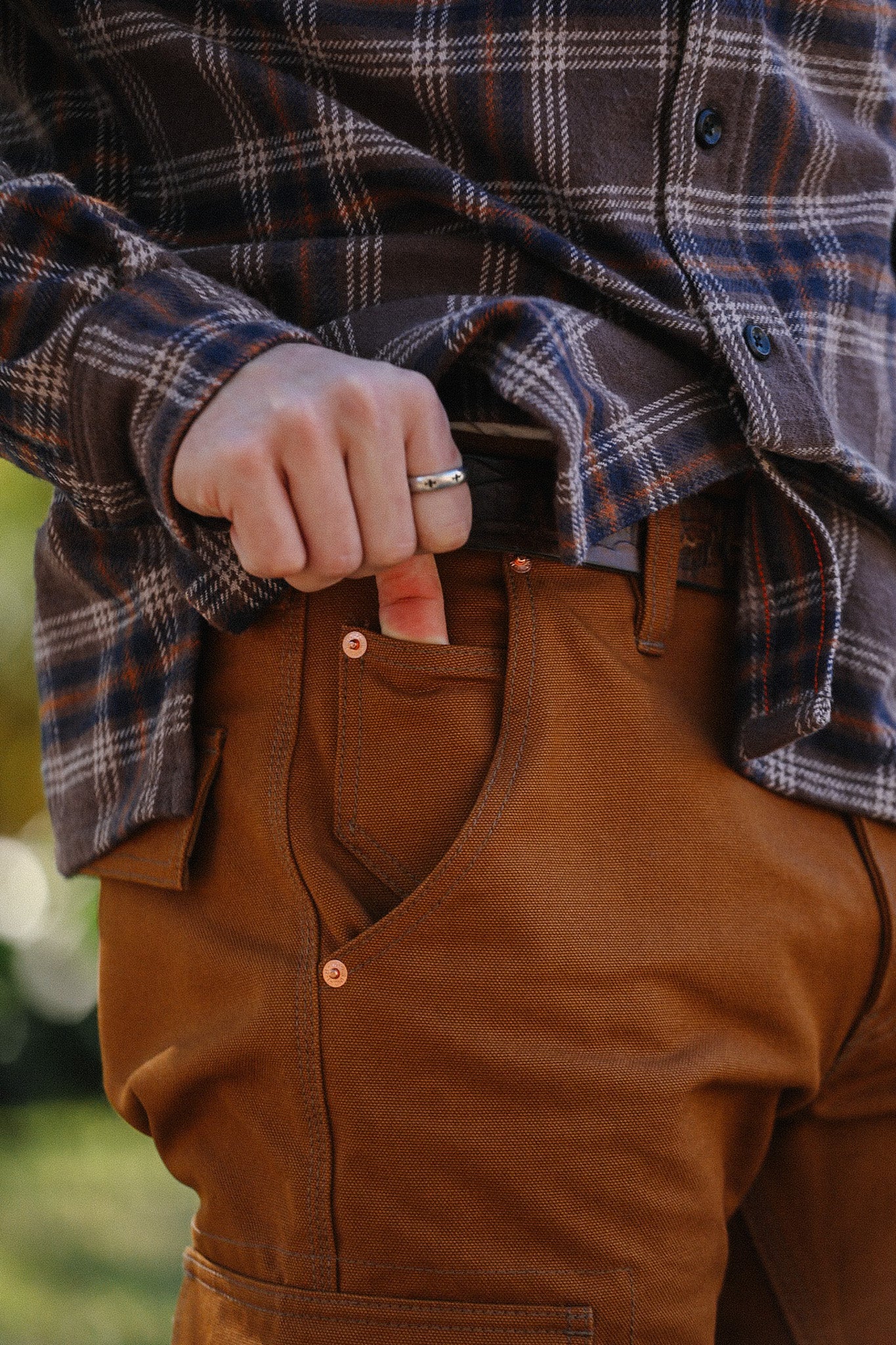 Close-up of person wearing brown work pants with hand in pocket and plaid shirt, showing ring detail