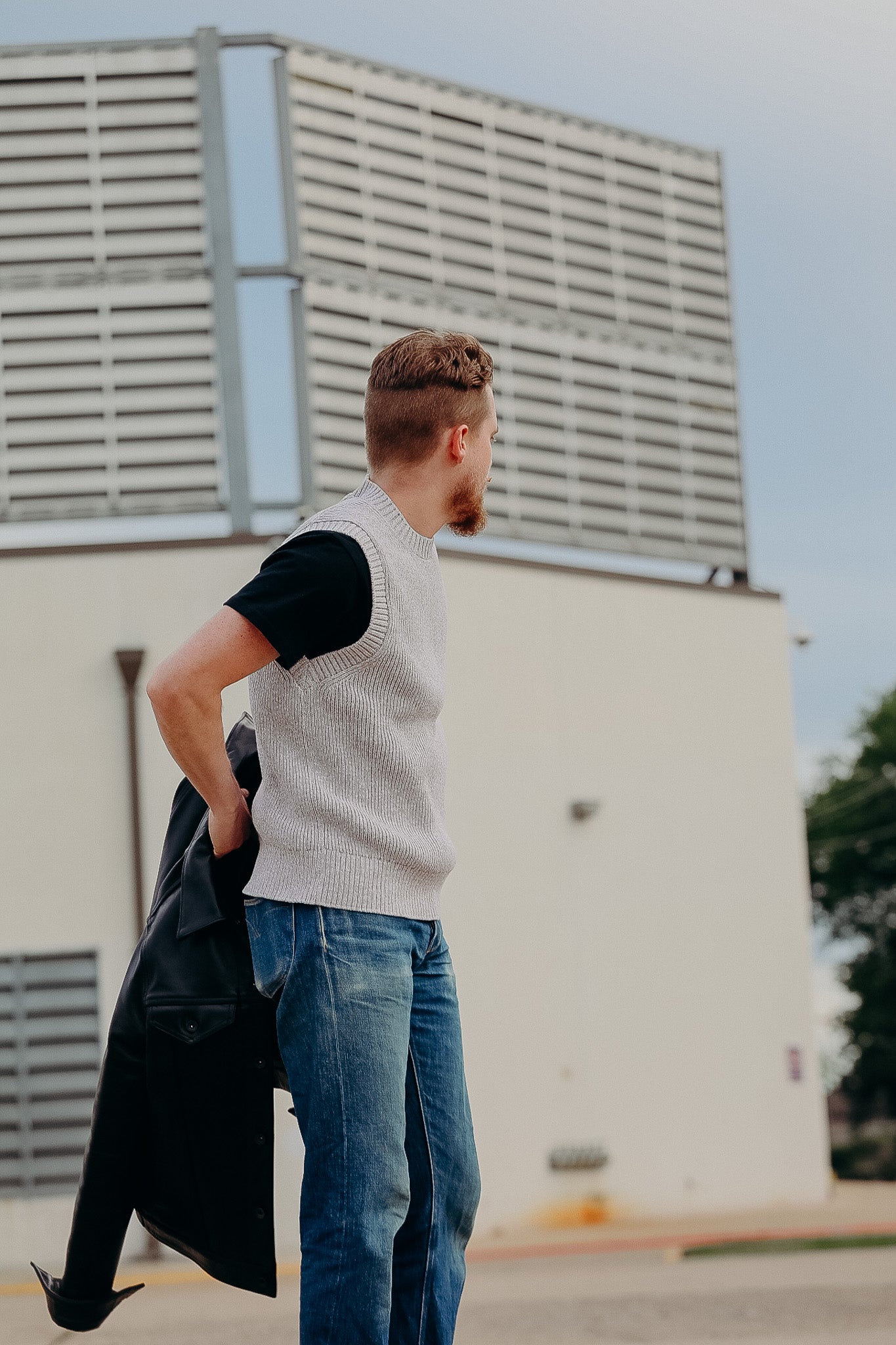 Man wearing gray knit vest and black t-shirt holding black leather jacket outdoors near modern building