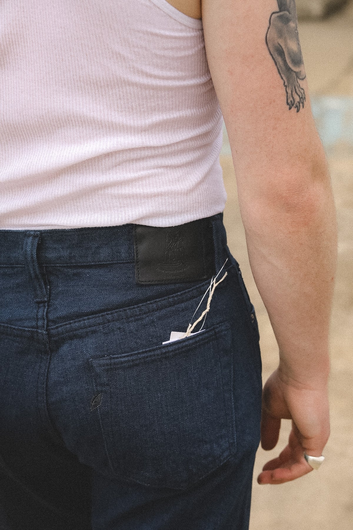 Close-up of the back of a person wearing dark blue denim jeans and a light pink ribbed tank top, with a tattoo visible on the upper arm.