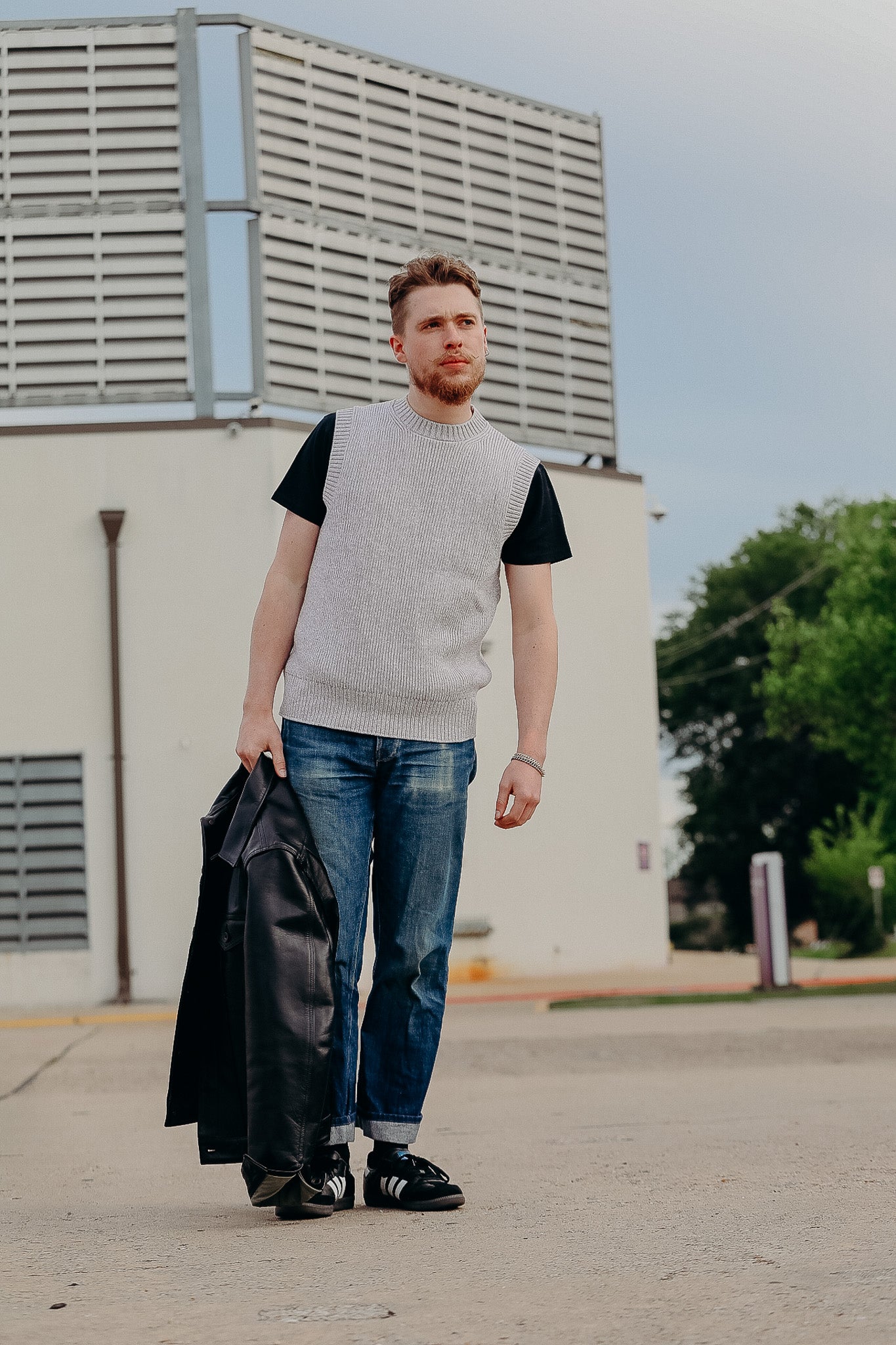 Man wearing gray knit vest over black t-shirt and jeans holding black leather jacket outdoors