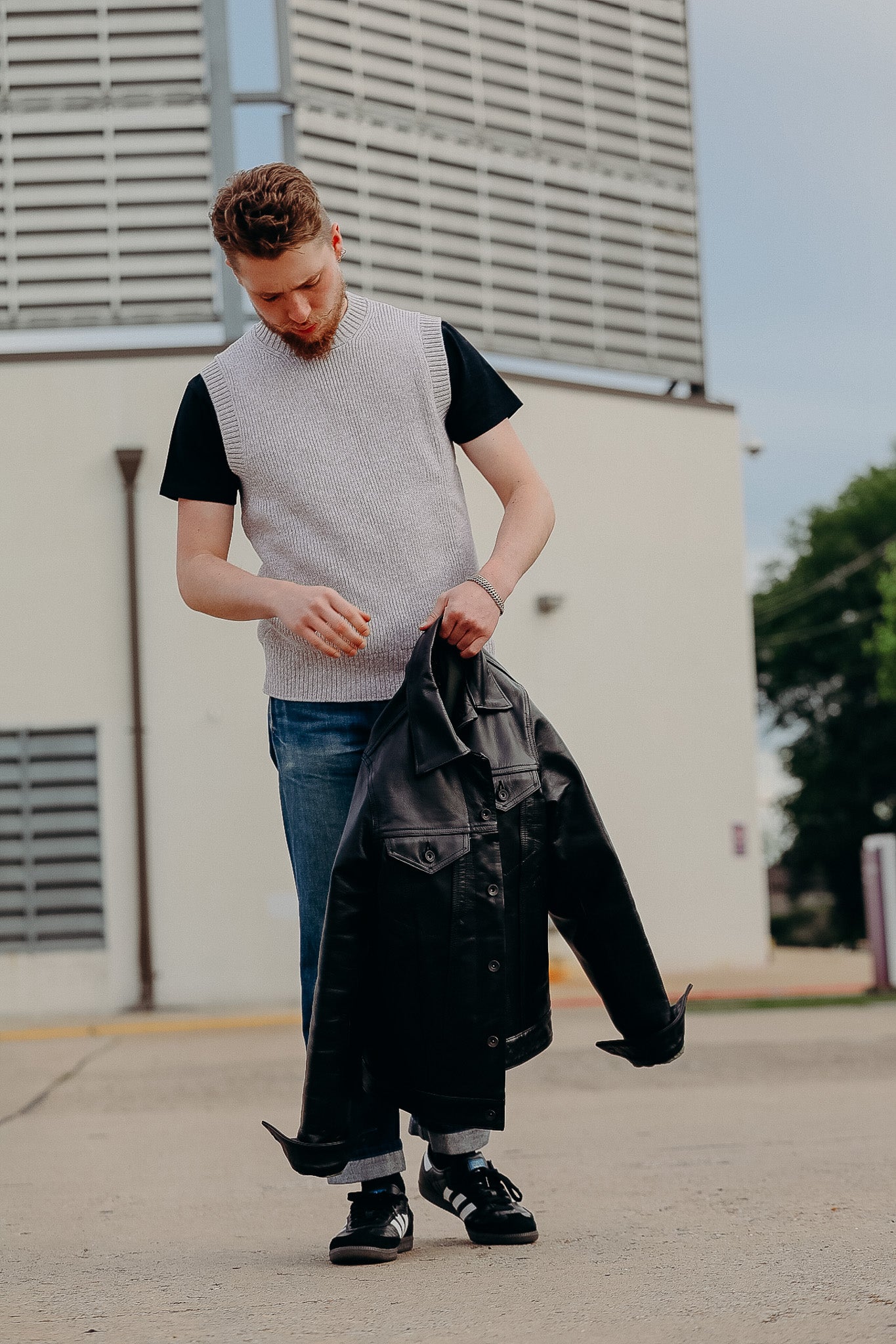 Man wearing a gray knit vest over black t-shirt, blue jeans, and black sneakers holding a black leather jacket outside