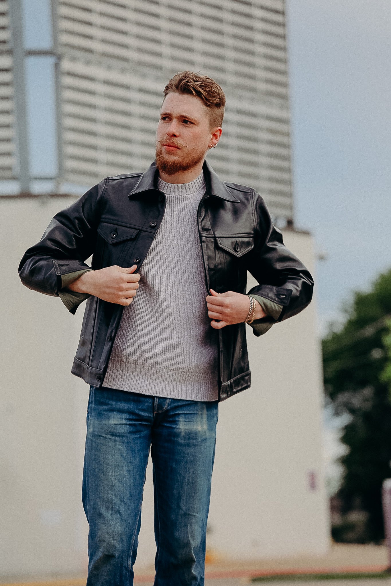 Man wearing black leather jacket over gray sweater and blue jeans, adjusting jacket outdoors near modern building