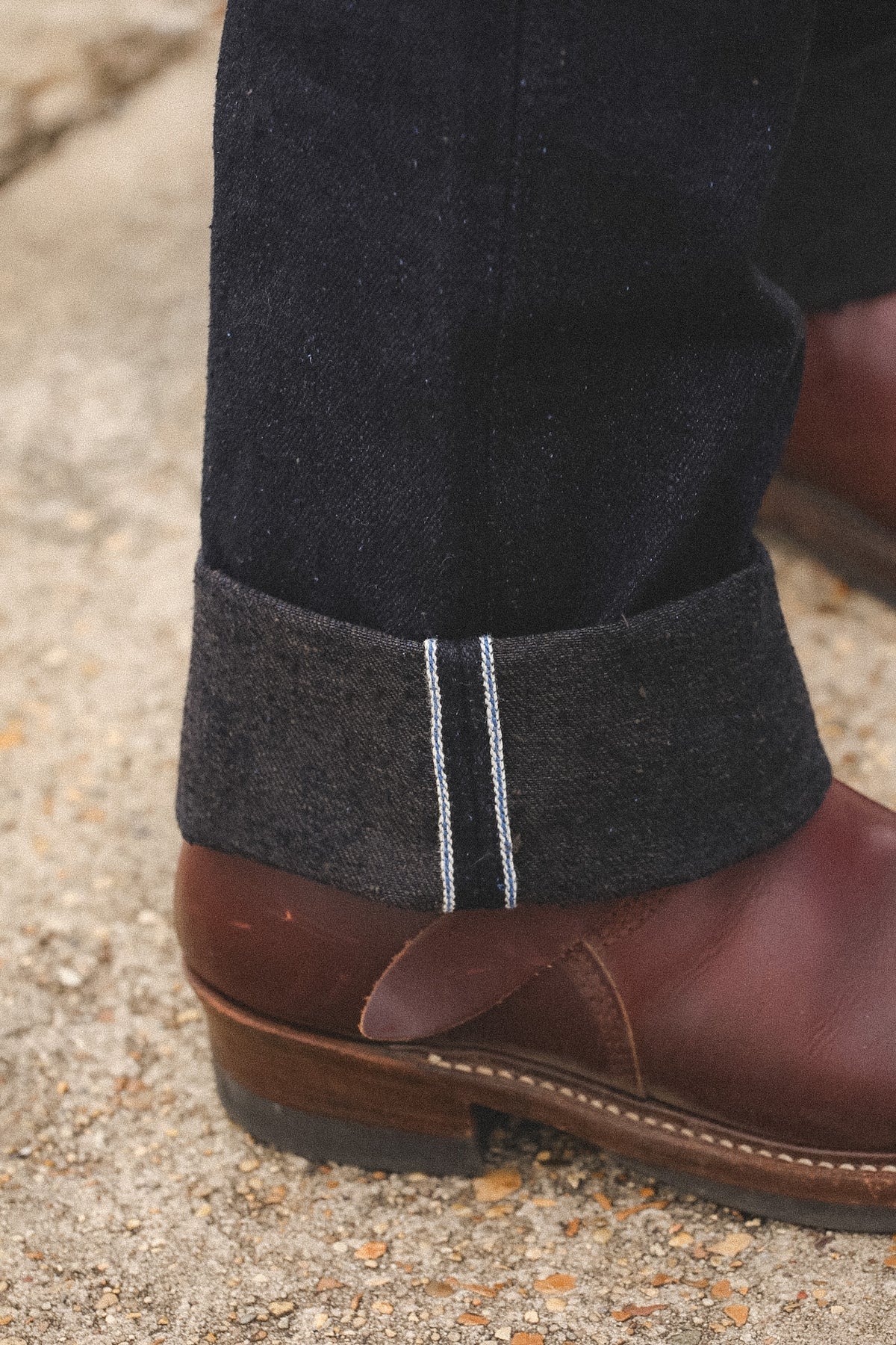 Close-up of a person's ankle wearing dark charcoal twill trousers with double white contrast stitching, rolled up to reveal a brown leather boot on a gravel surface.
