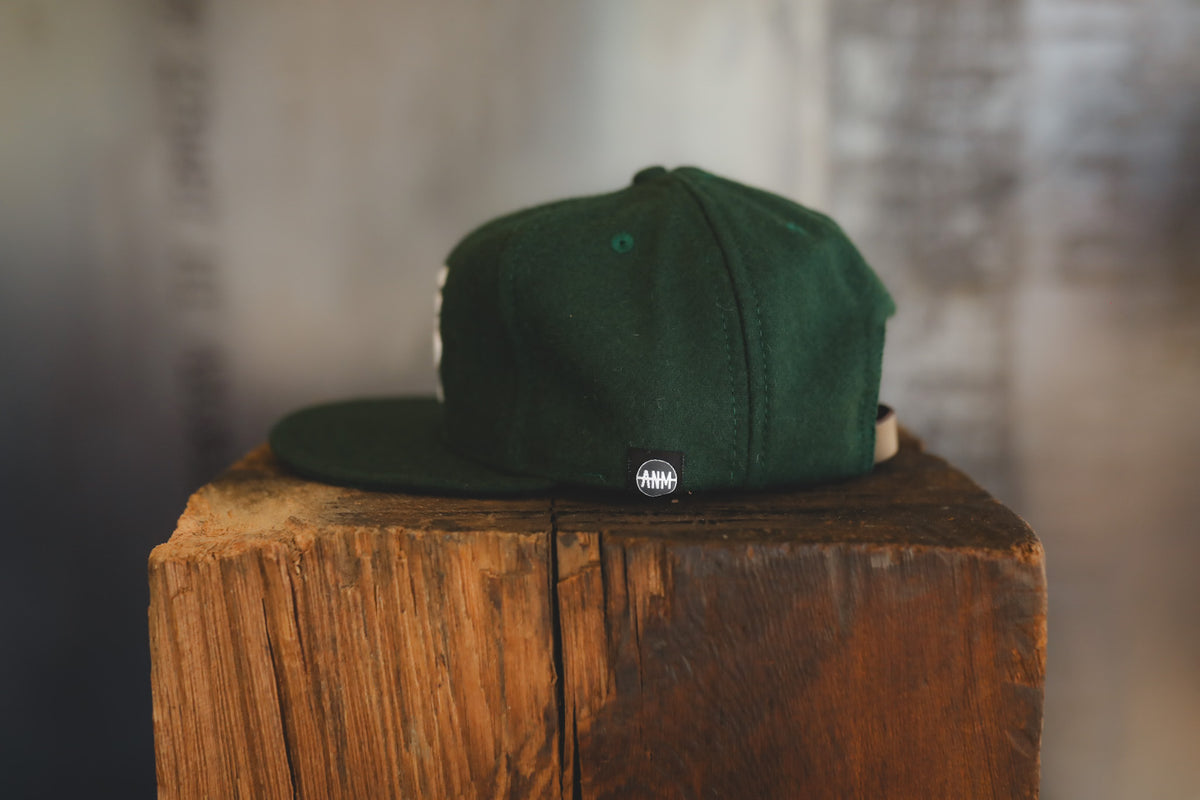 A dark green snapback cap with a small circular black and white logo on the side, displayed on a rustic wooden block against a blurred gray background.