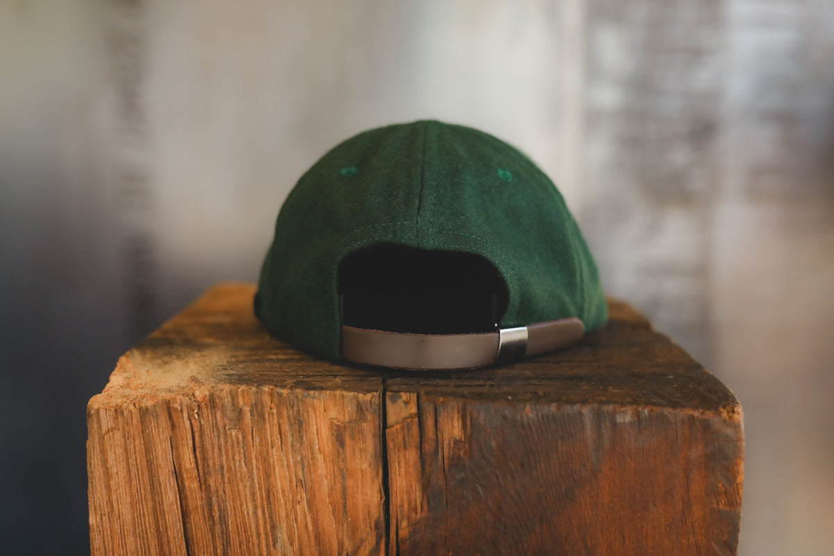 A green baseball cap viewed from the back, resting on a rustic wooden block with a softly blurred gray background.