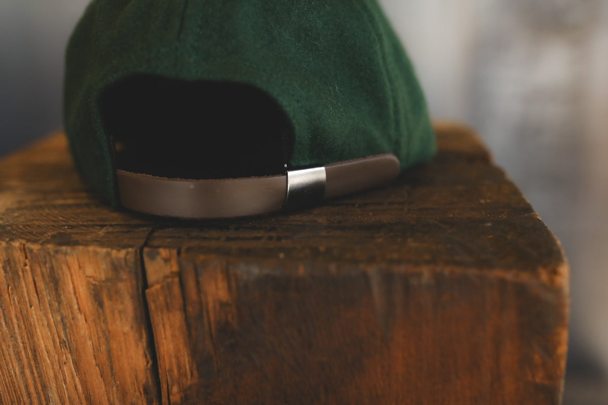 A close-up shot of the back of a dark green cap resting on a rustic wooden block, showing a metallic silver adjustment strap.