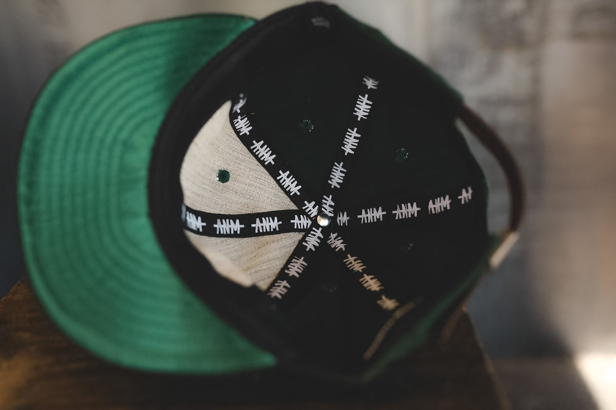 A close-up of a snapback cap with a green brim, black crown, and a white front panel embroidered with white runic symbols.