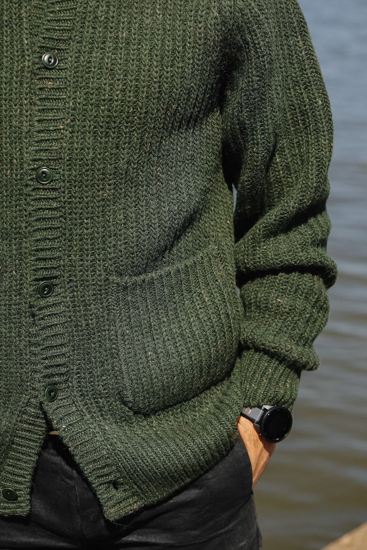 Close-up of a person wearing an olive green ribbed knit cardigan with front buttons and a chest pocket, standing outdoors near water.