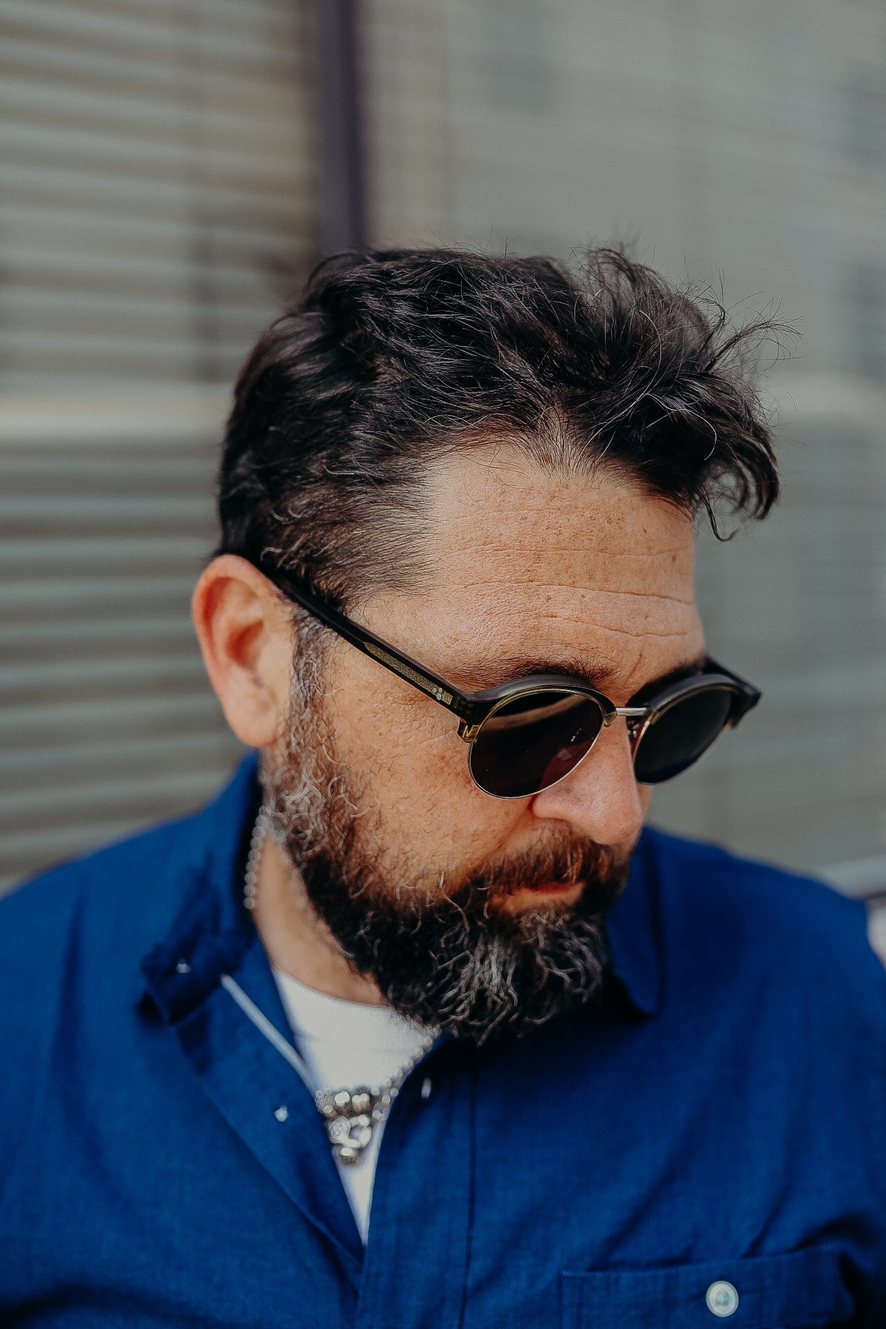 Man with dark hair and beard wearing black sunglasses and a blue shirt against a neutral background