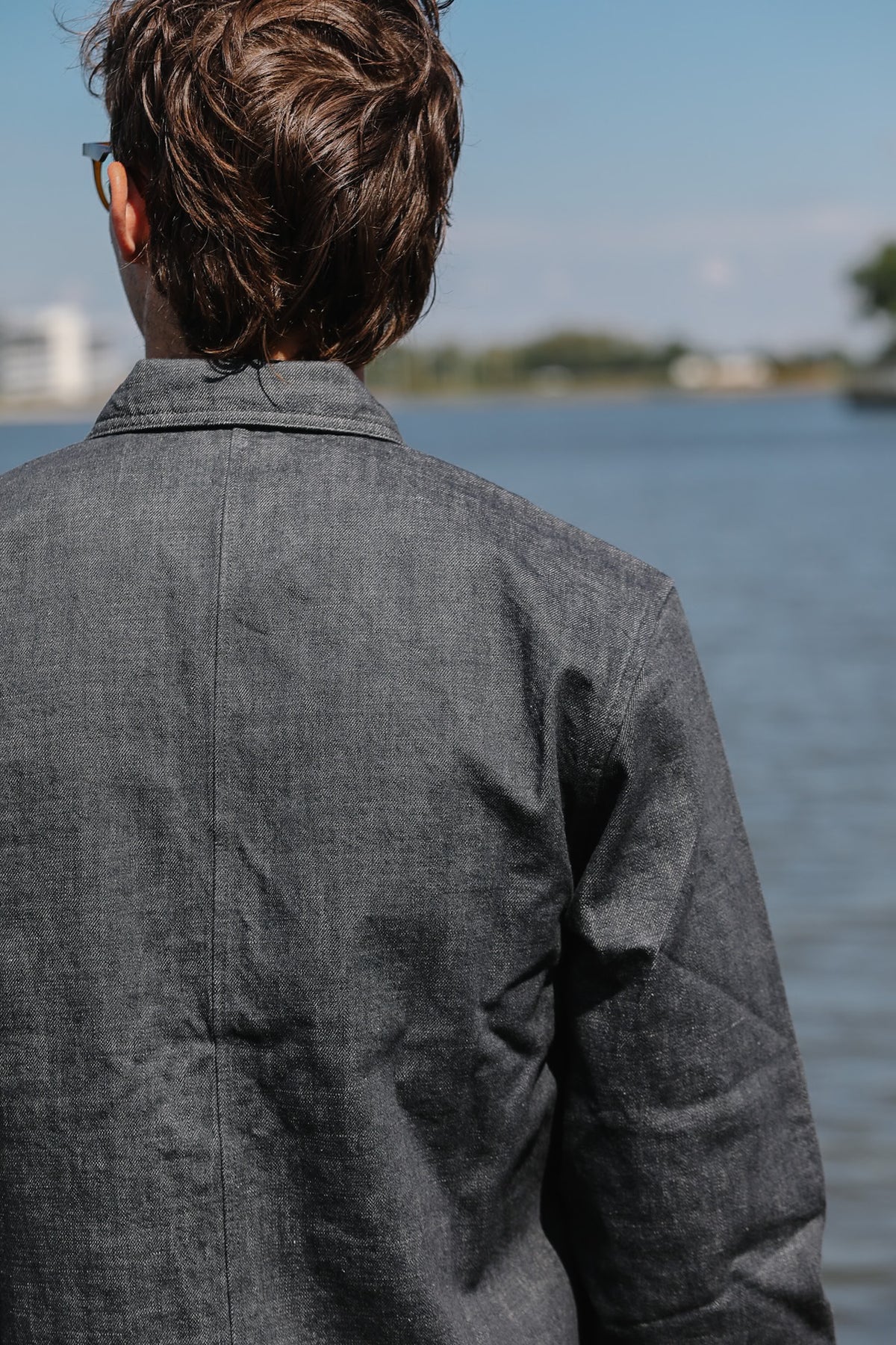 A man viewed from behind wearing a dark gray textured linen shirt, standing outdoors with a body of water and distant shoreline in the background.