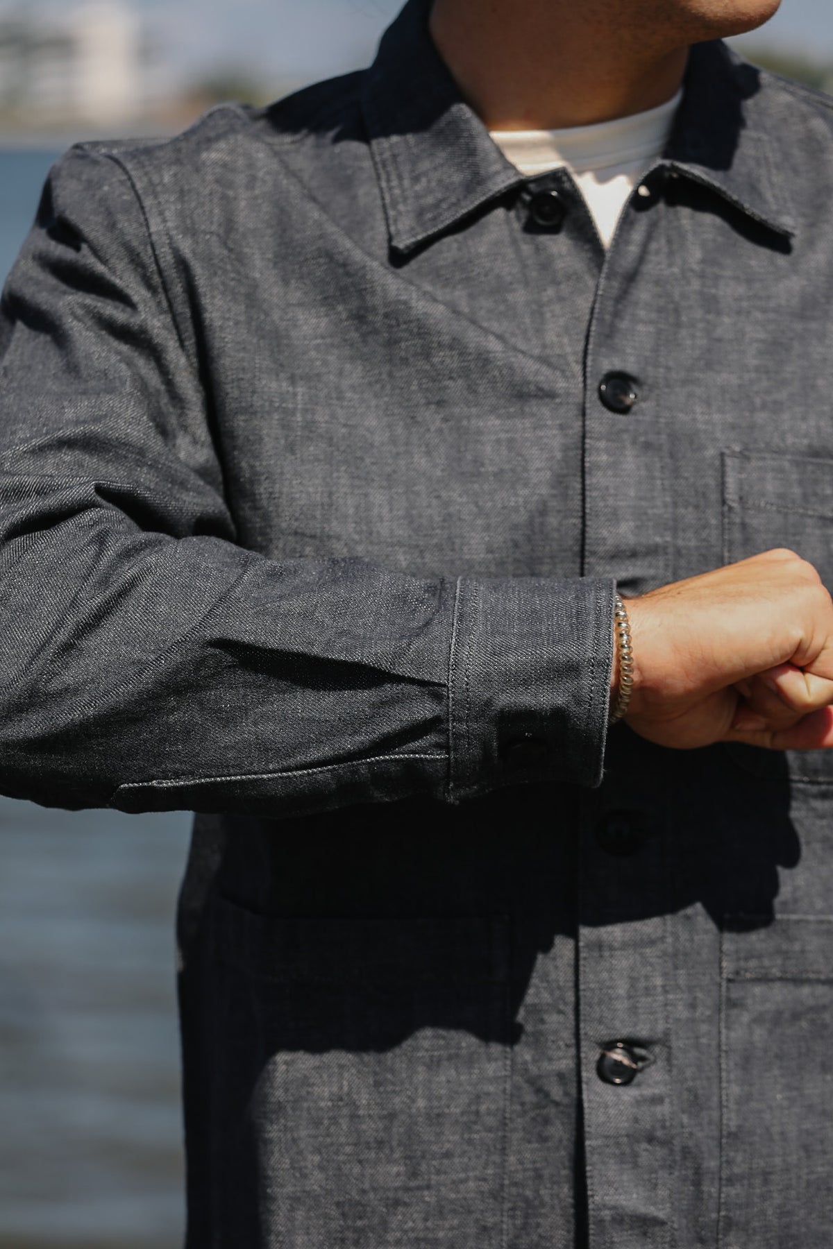 A close-up of a person wearing a dark gray textured overshirt or jacket with black buttons, layered over a white undershirt, with water and a blurred outdoor background.
