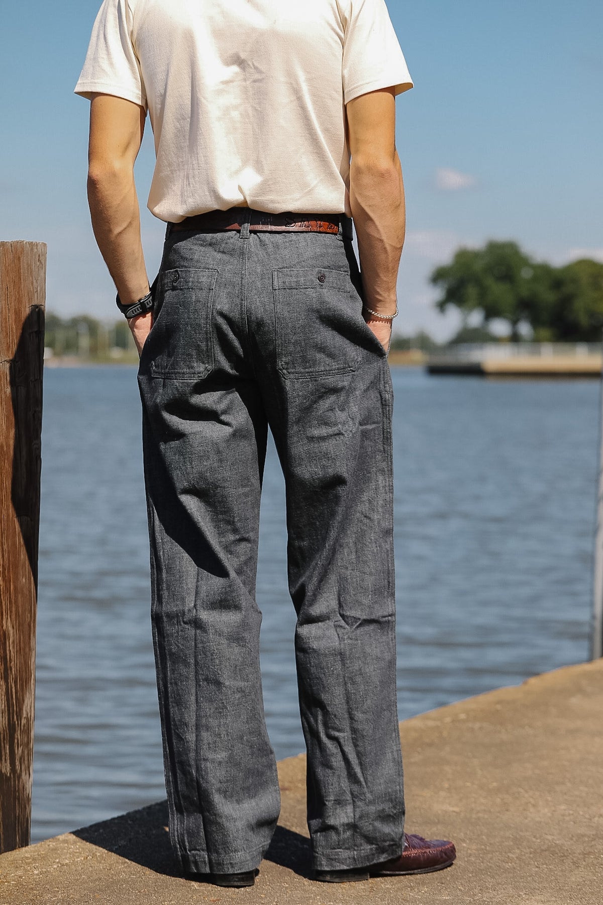 A person standing outdoors by a body of water, shown from the back wearing dark gray wide-leg trousers, a cream t-shirt, and brown shoes.