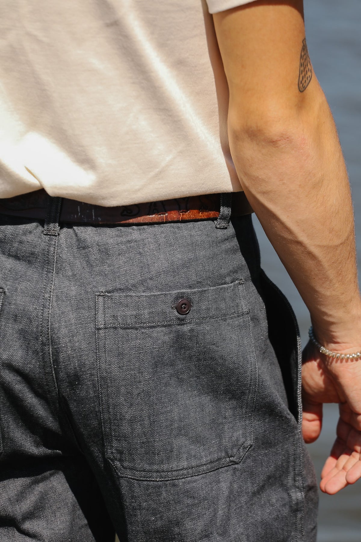 Close-up back view of a person wearing dark gray textured denim work pants with a back pocket and button, tucked into a cream t-shirt.