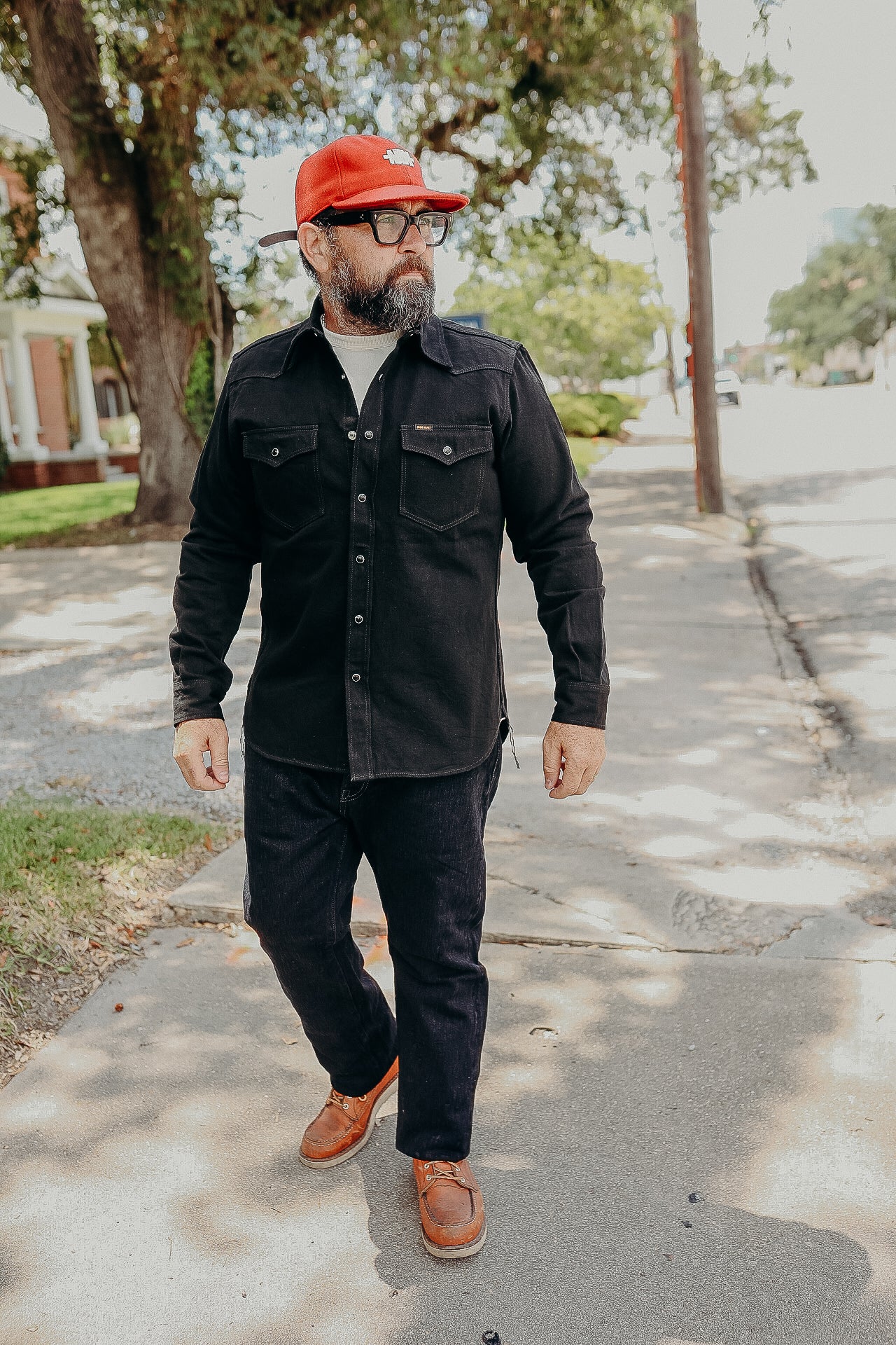 Man wearing black work shirt, dark jeans, red cap, and brown leather boots walking on sidewalk under trees