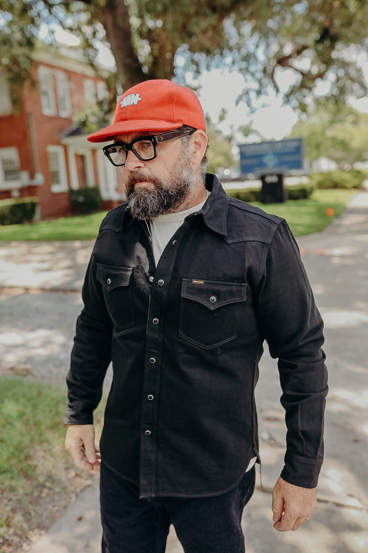 Bearded man wearing black work shirt and red cap with glasses outdoors near a brick building