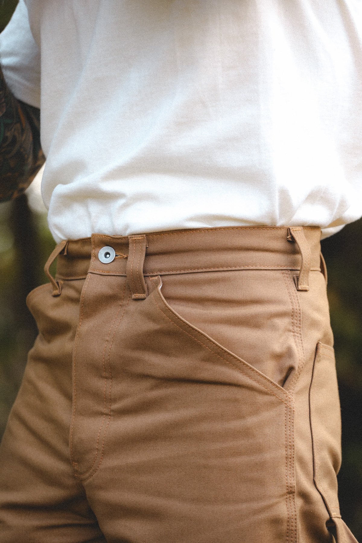 Close-up of a person wearing tan chinos with a white t-shirt, showing the waistband, belt loops, button, and front pocket details.