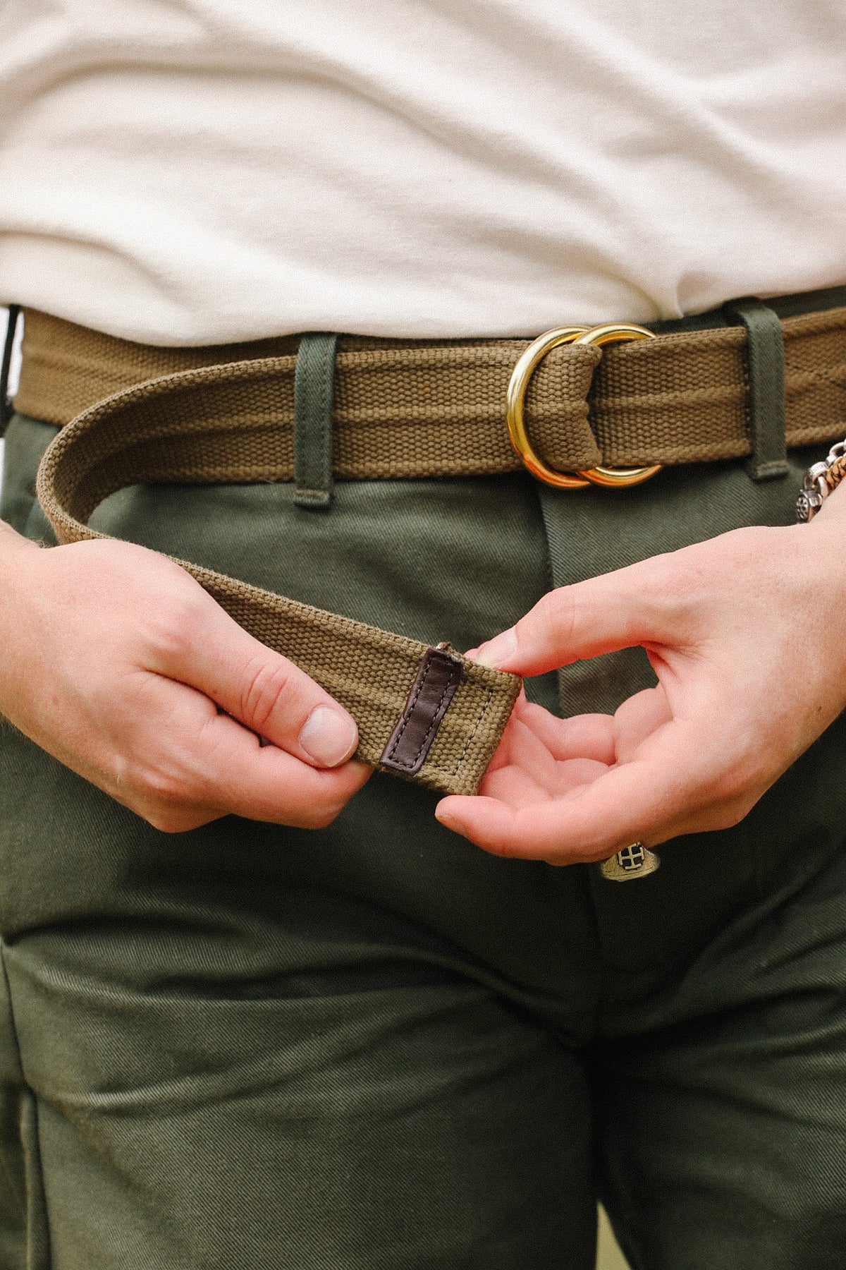 Close-up of hands adjusting olive green canvas belt with double gold rings worn with green pants and white shirt