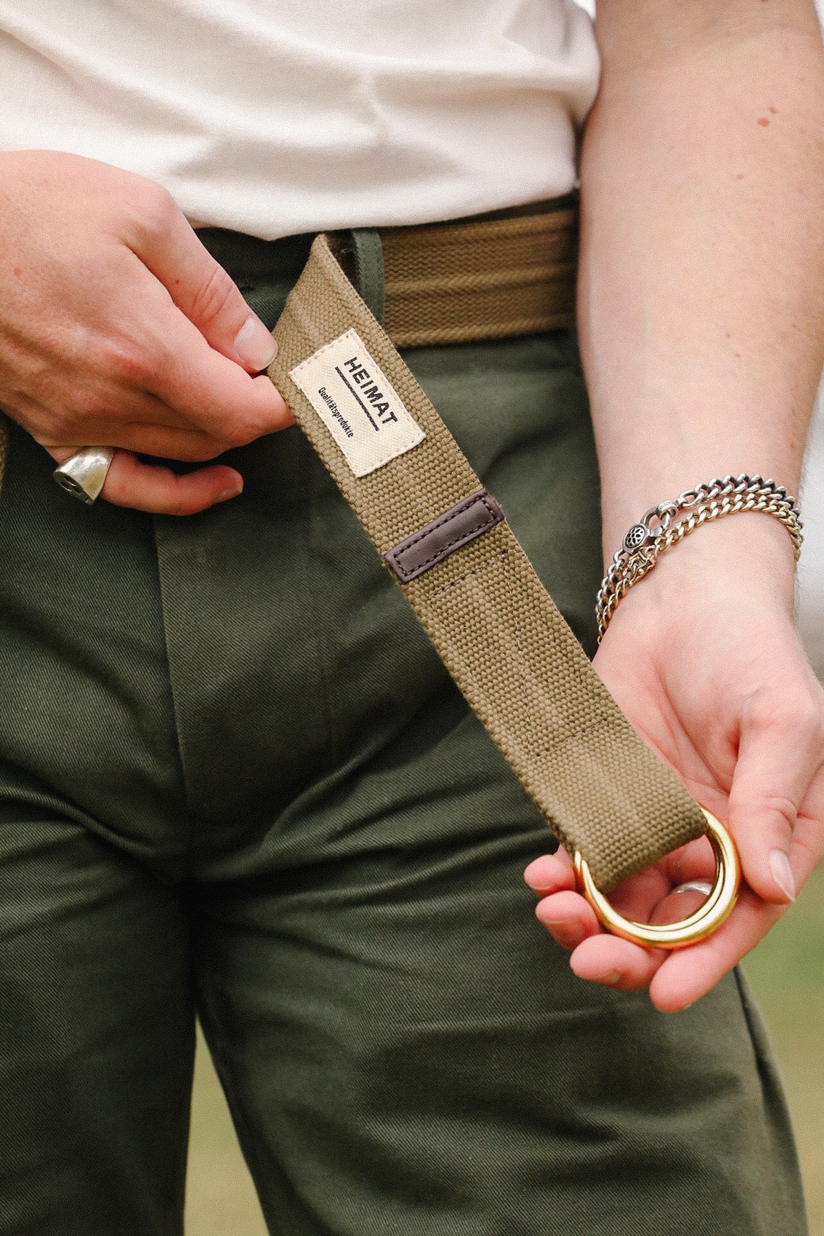 Close-up of a person wearing olive green pants and holding a khaki canvas belt with brass rings and a Heimat label