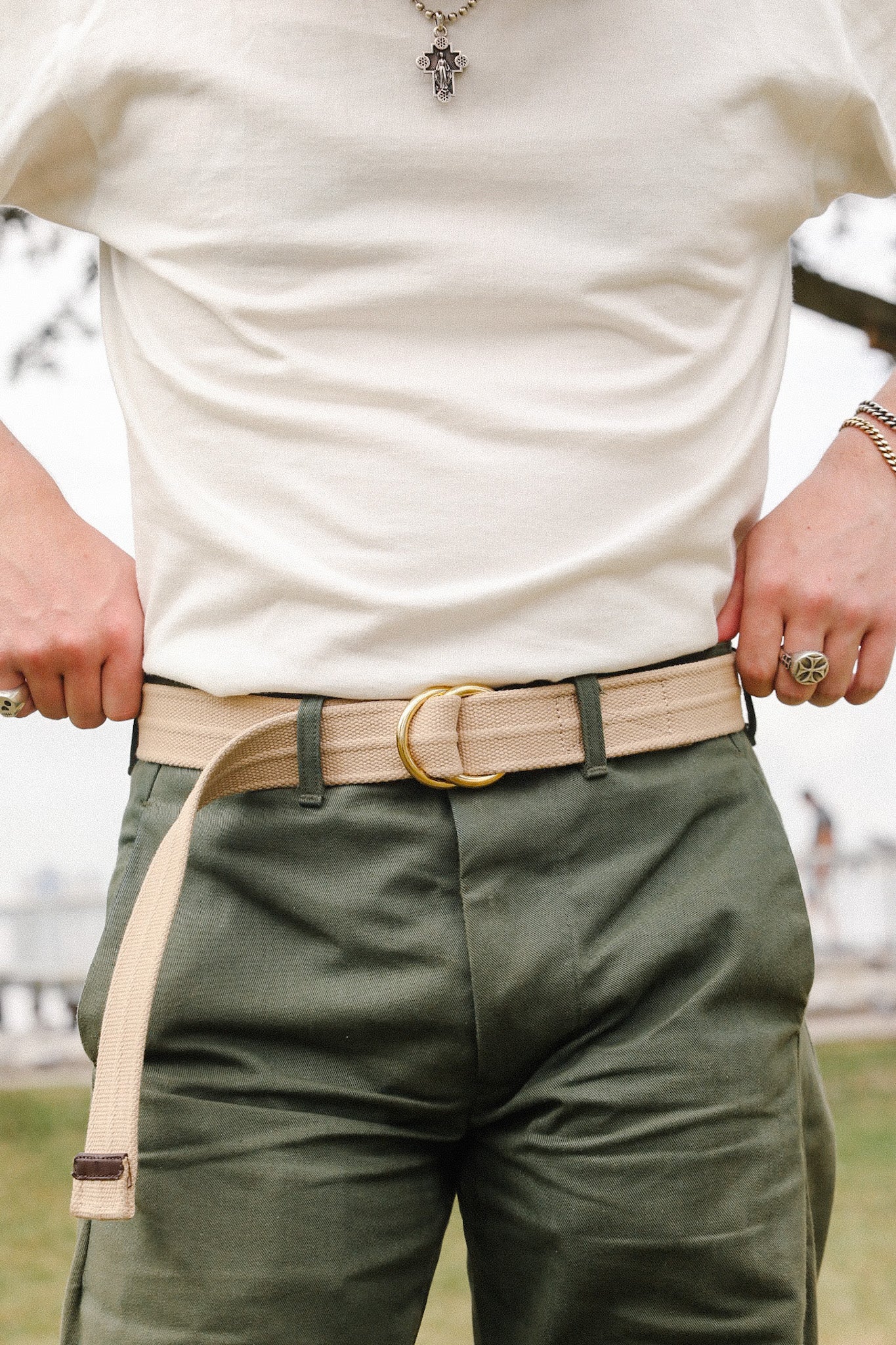 Person wearing beige canvas belt with gold buckle, olive green pants, white shirt, and silver cross necklace