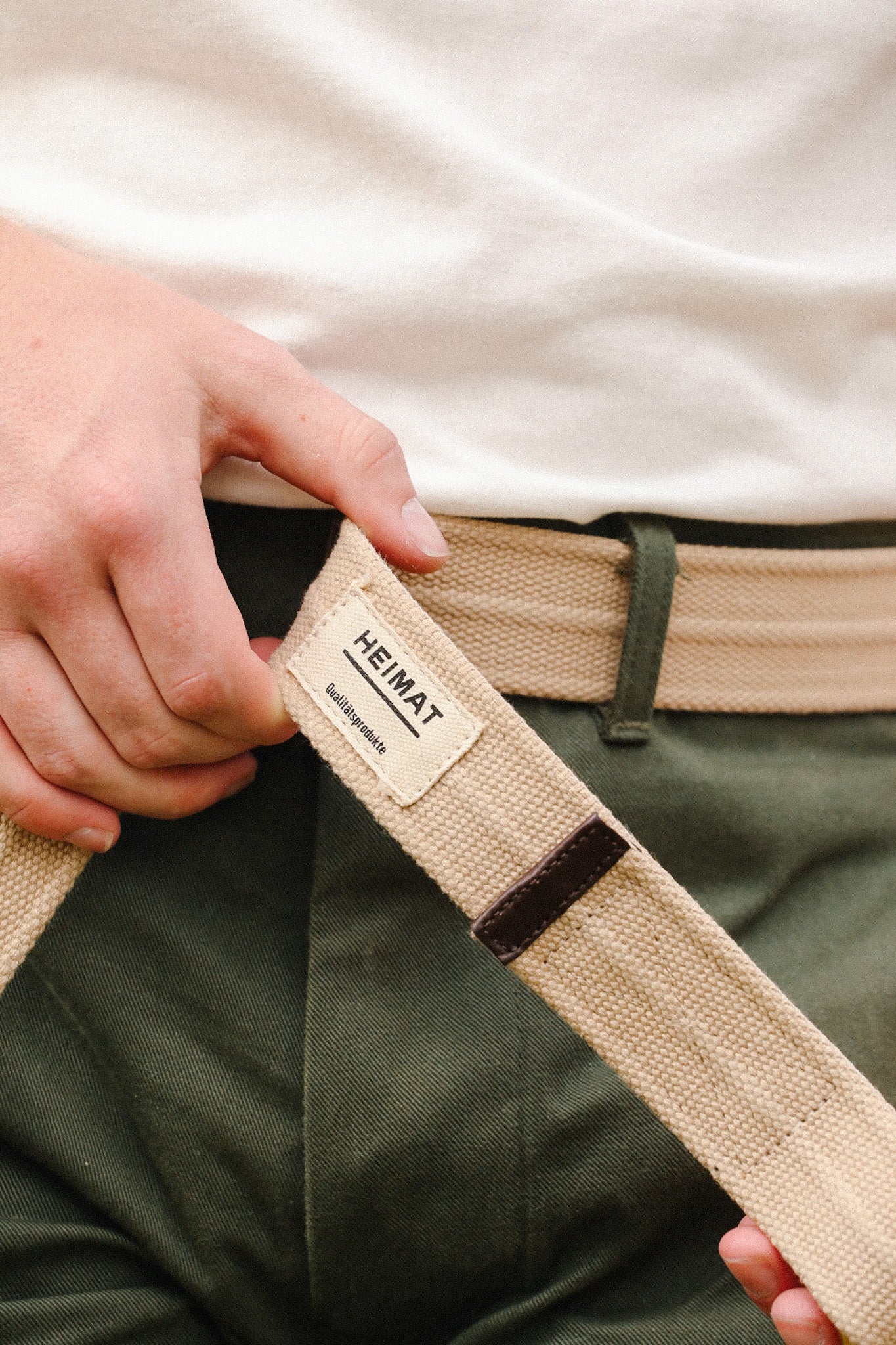 Close-up of hand holding beige canvas belt with Heimat label, worn with green pants and white shirt