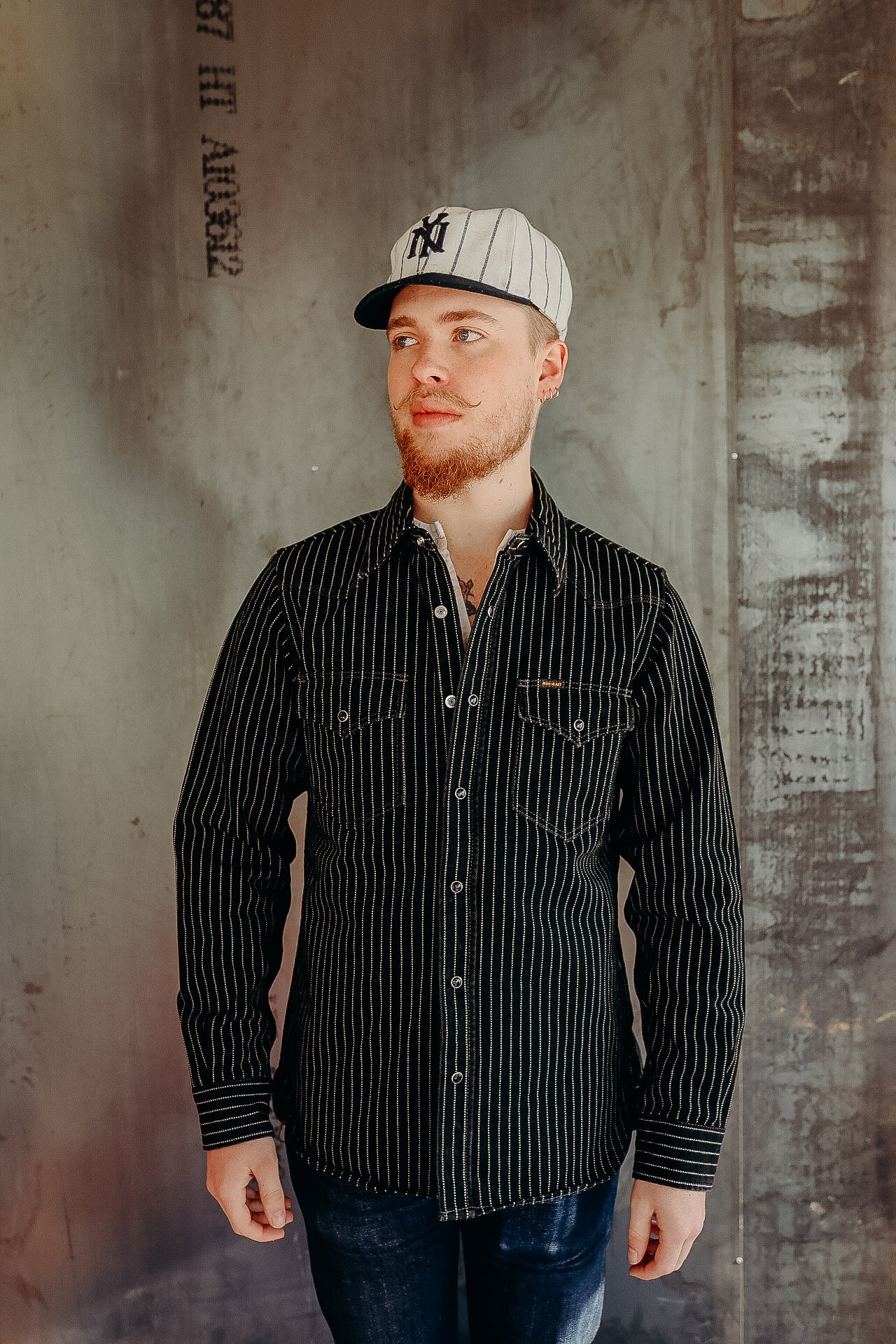 Young man wearing black striped work shirt and white pinstripe baseball cap standing against industrial concrete wall